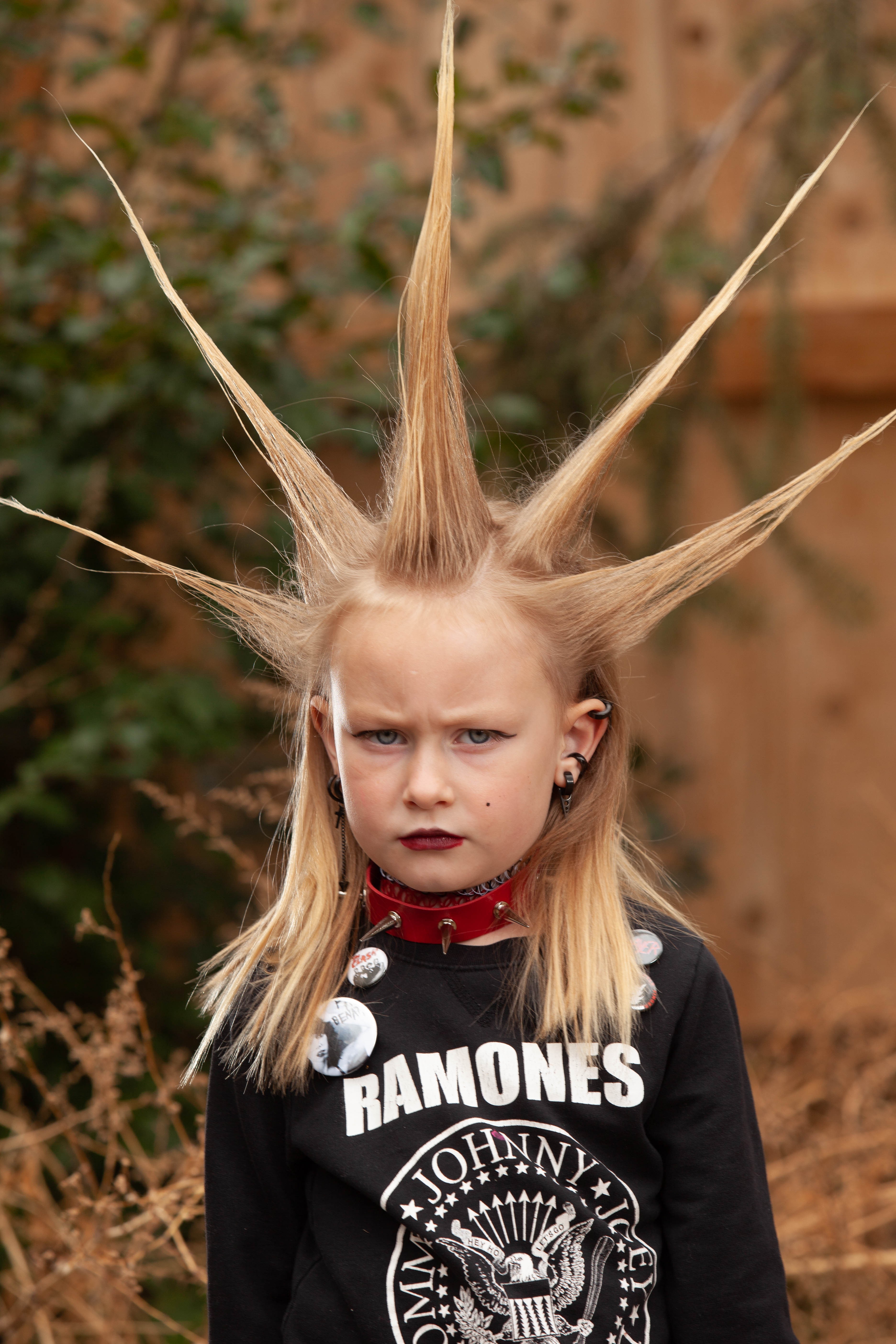 Young girl with spiky hair standing outdoors, wearing a Ramones shirt and a red choker, with a serious expression.