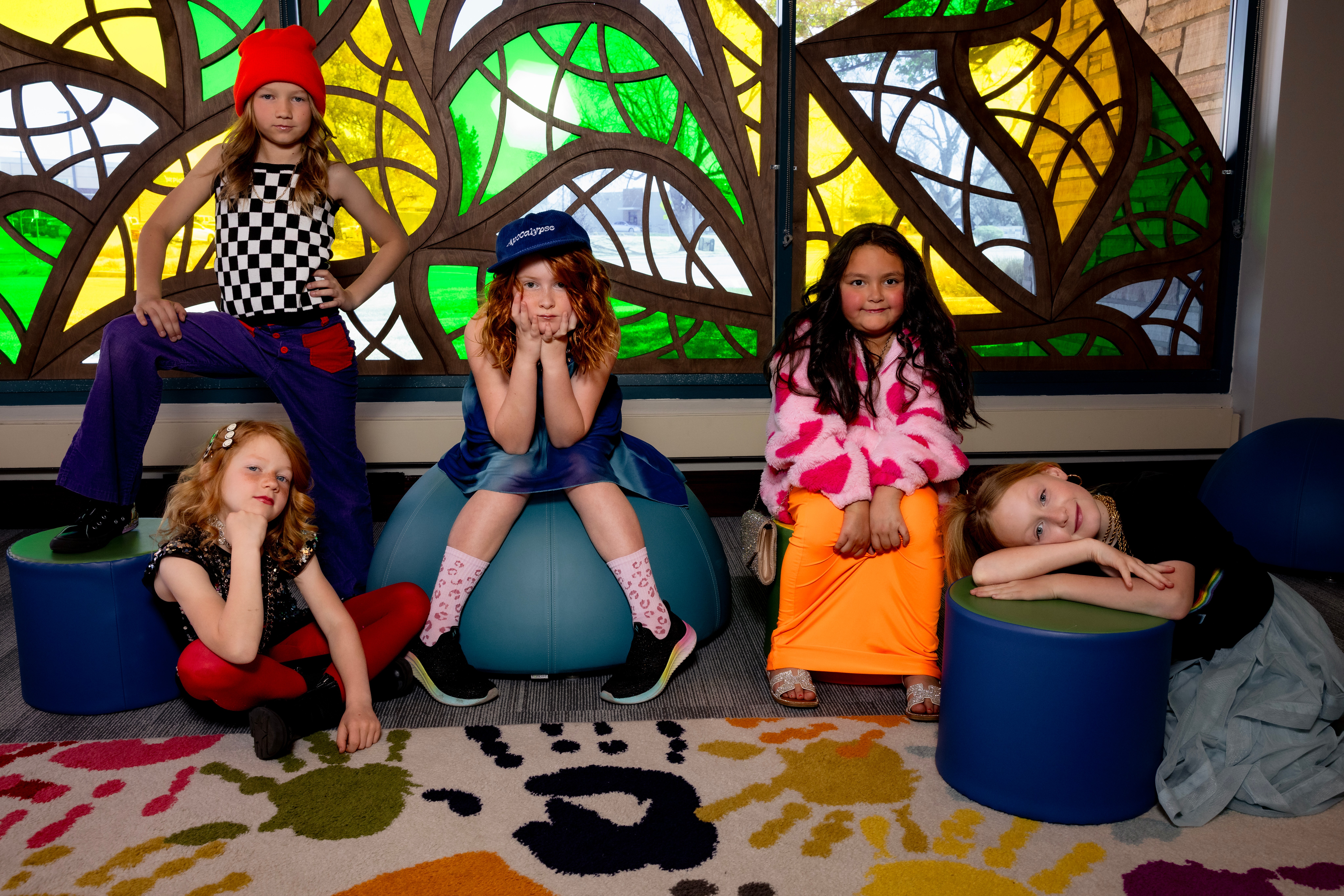 Five children sitting and lying on colorful cushions in front of stained glass windows, smiling and posing. Word art on carpet.