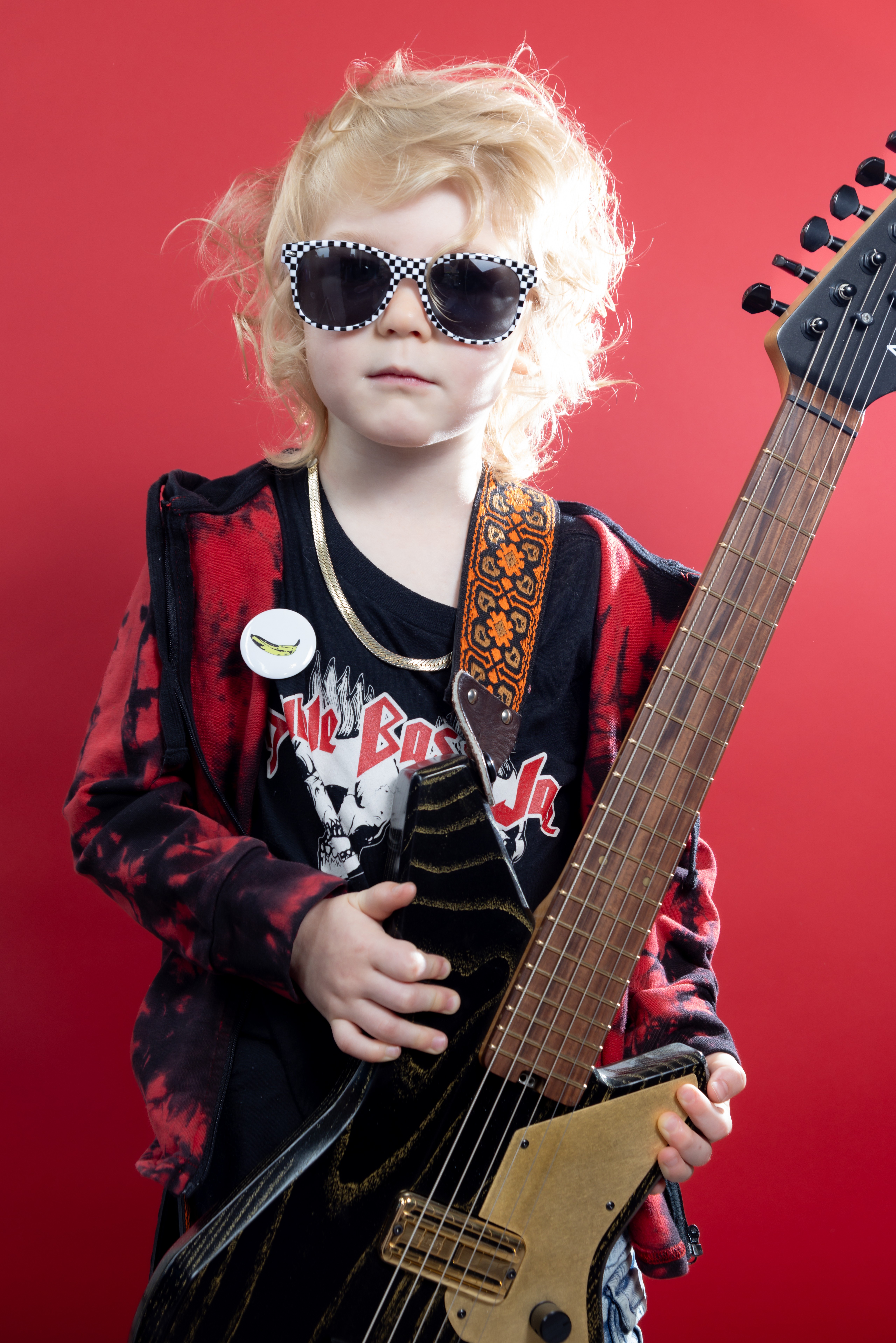 Child with curly blonde hair wearing sunglasses, a black and red jacket, holding an electric guitar against a red background.