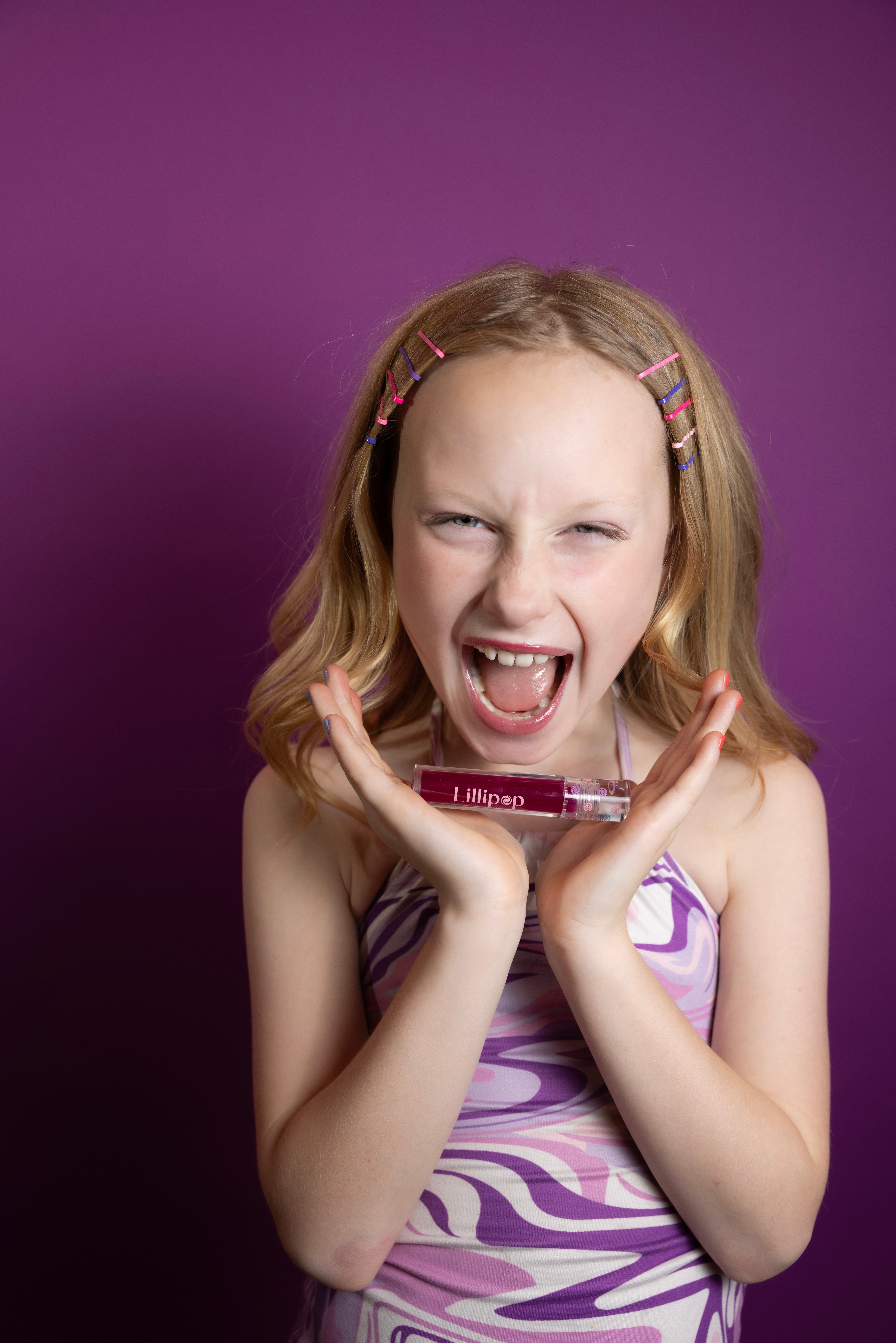 Young girl with blonde hair and colorful hair clips, smiling and holding a lipstick, against a purple background.
