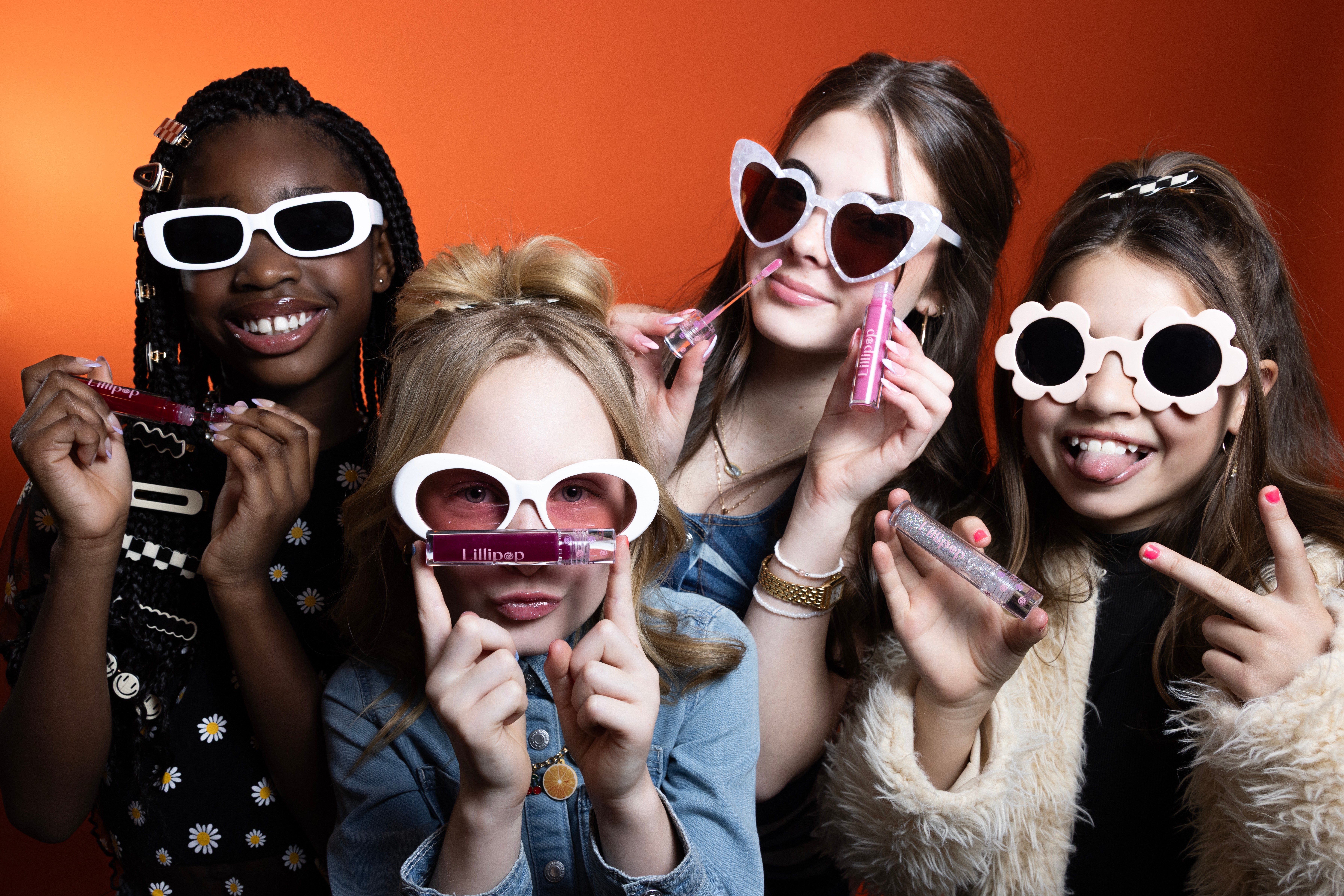 Four people wearing large sunglasses and holding makeup brushes, smiling against a red background.