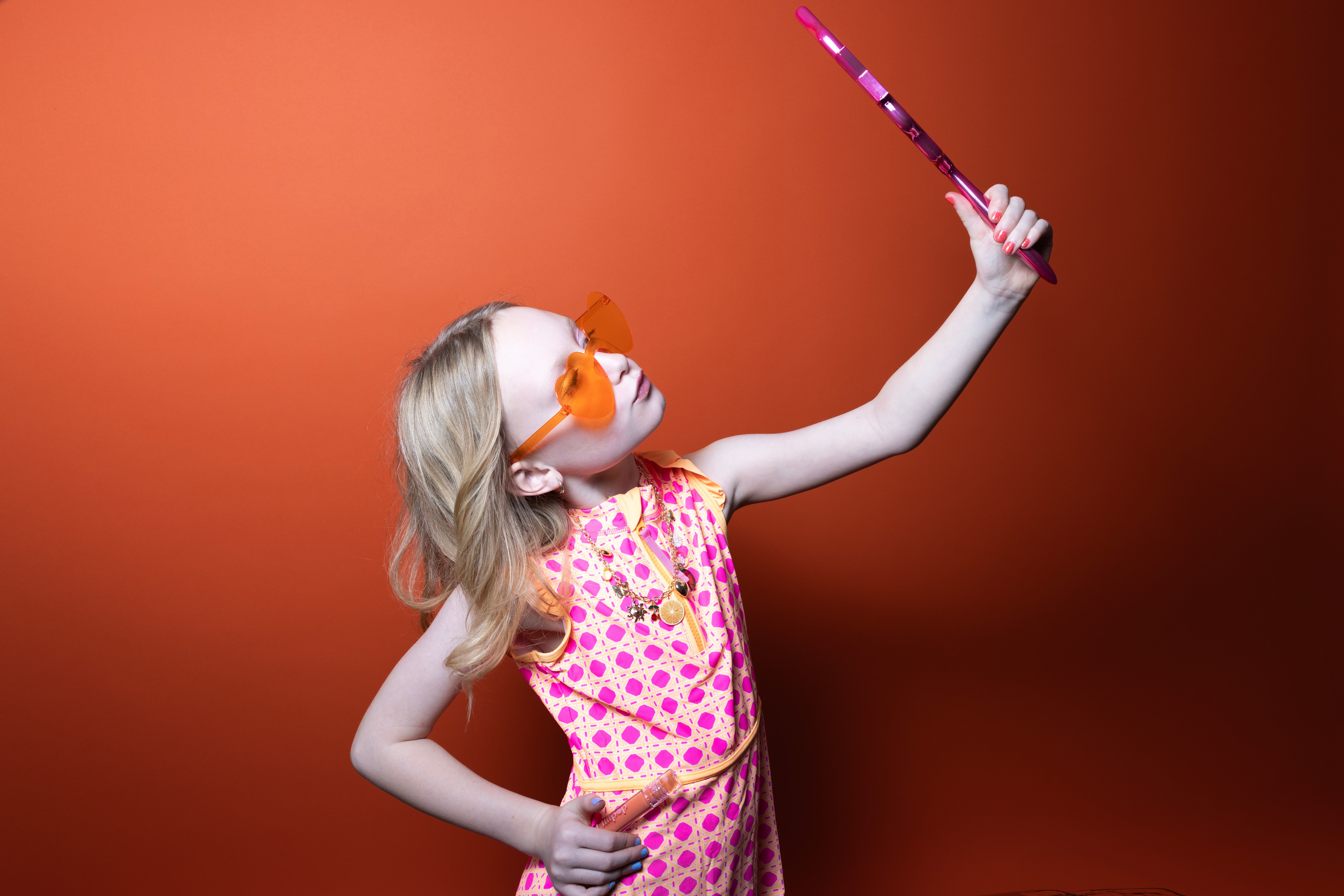 Young girl with blonde hair wearing sunglasses and a pink polka dot dress, holding a selfie stick against an orange background.
