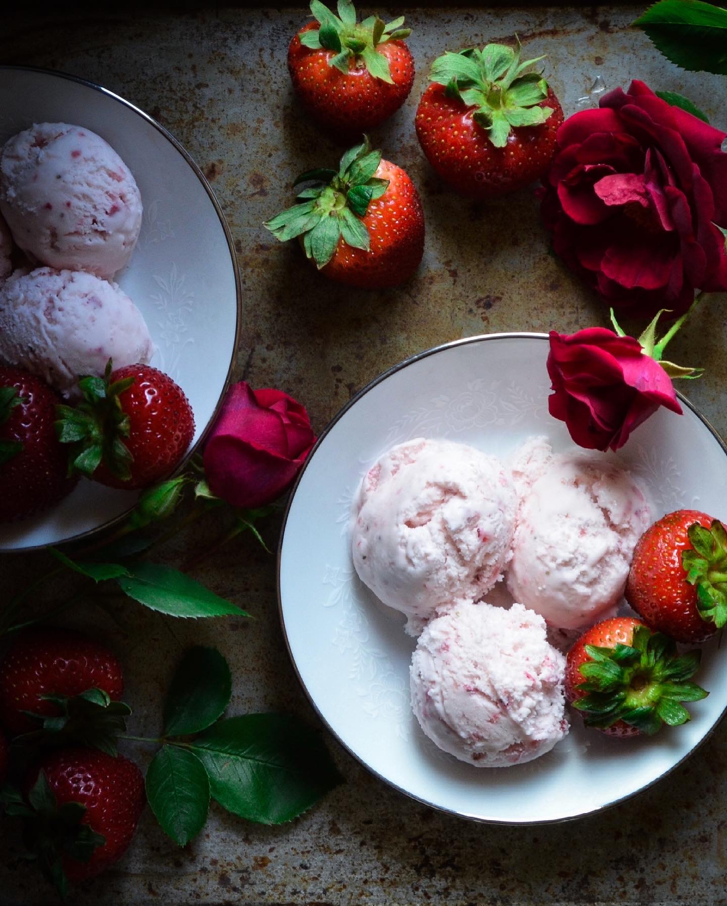 Strawberries, roses, and scoops of ice cream on plates and a surface, with some strawberries and roses nearby.
