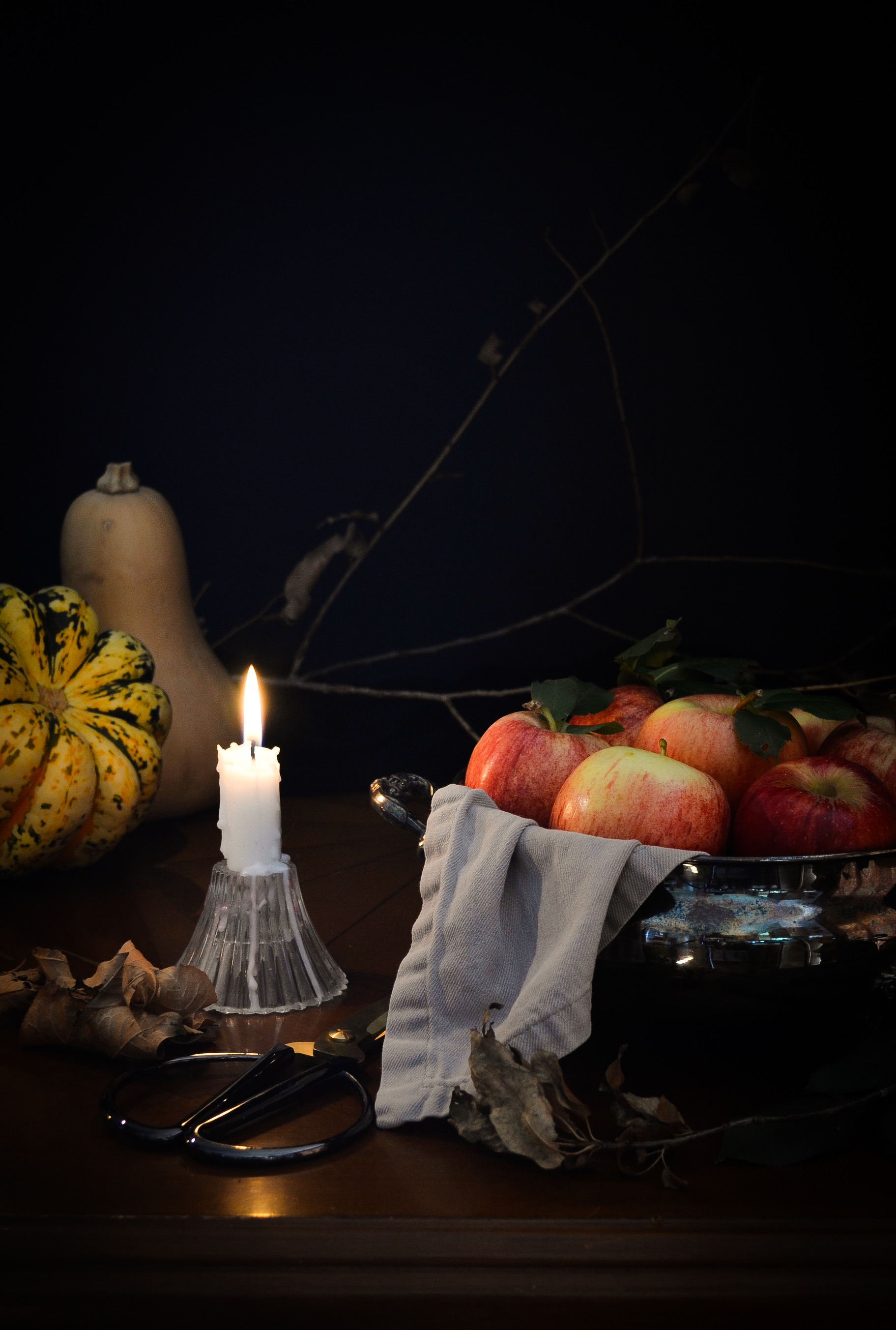A lit candle, apples in a bowl, a yellow gourd, and a pair of glasses on a dark table with a black background.