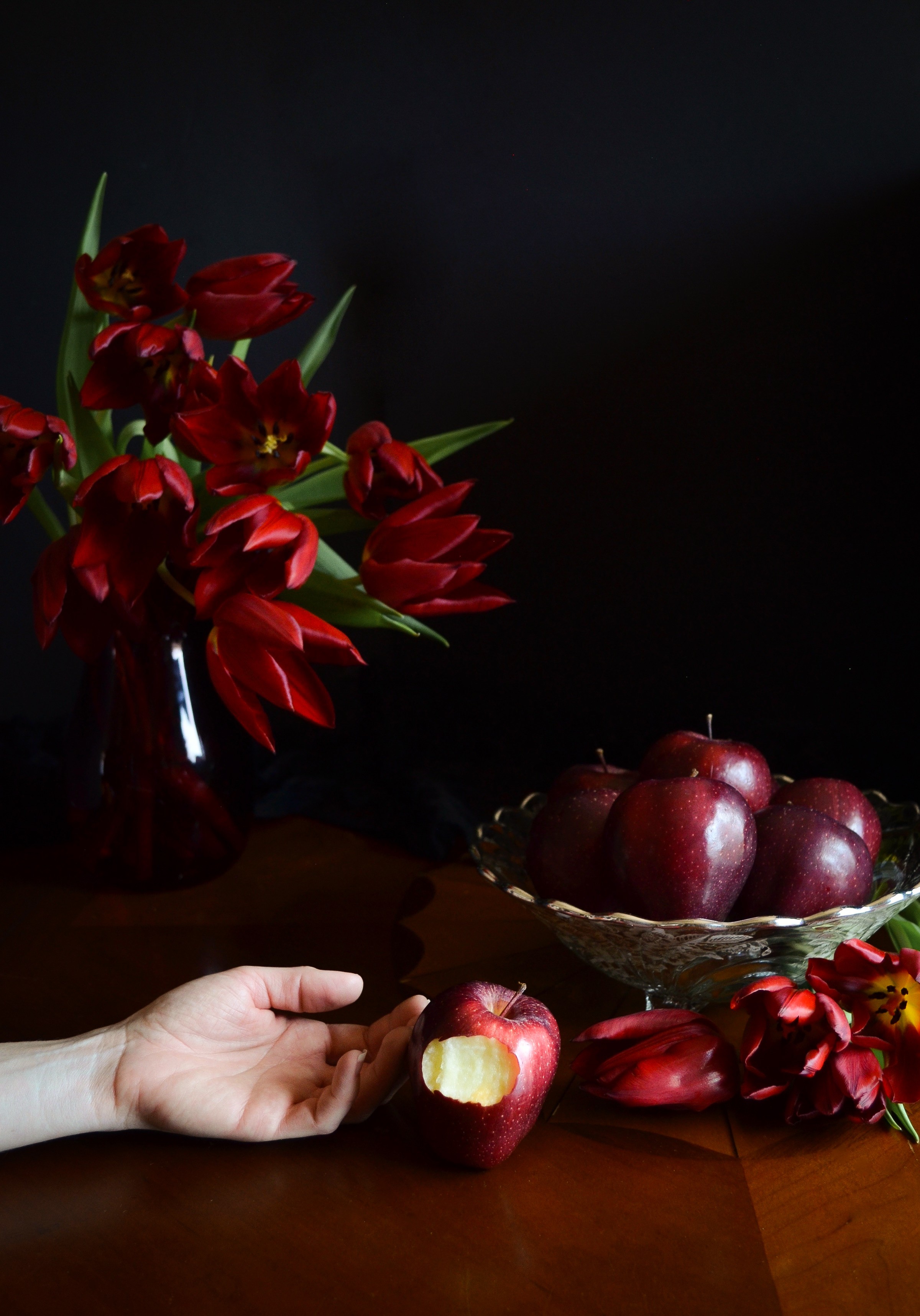 Hand holding a partially eaten red apple on a wooden table with red flowers and apples in the background.