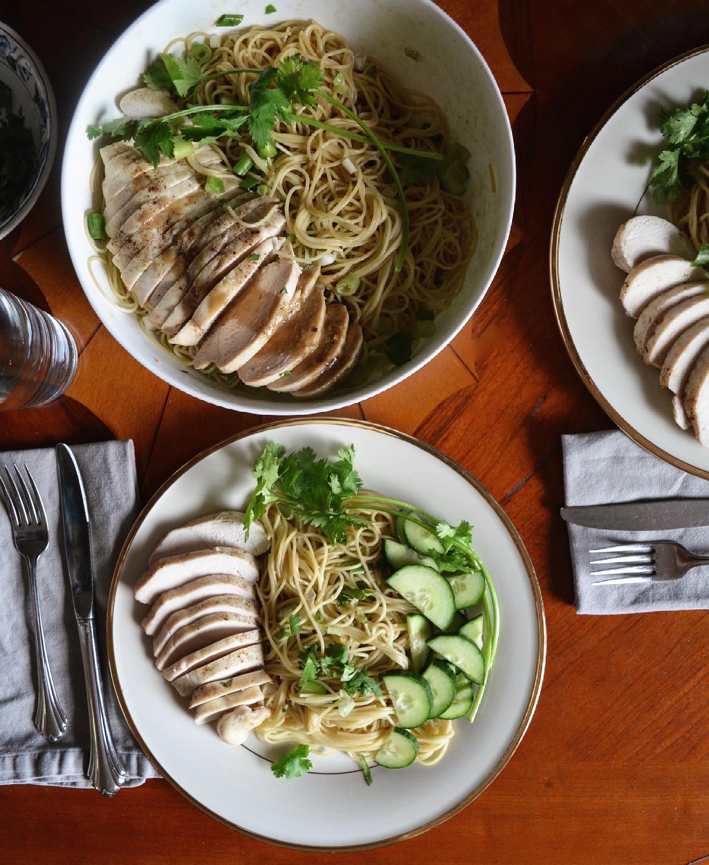 Bowl of noodles with sliced meat, herbs, and vegetables, along with two plates of sliced meat and vegetables on a wooden table.