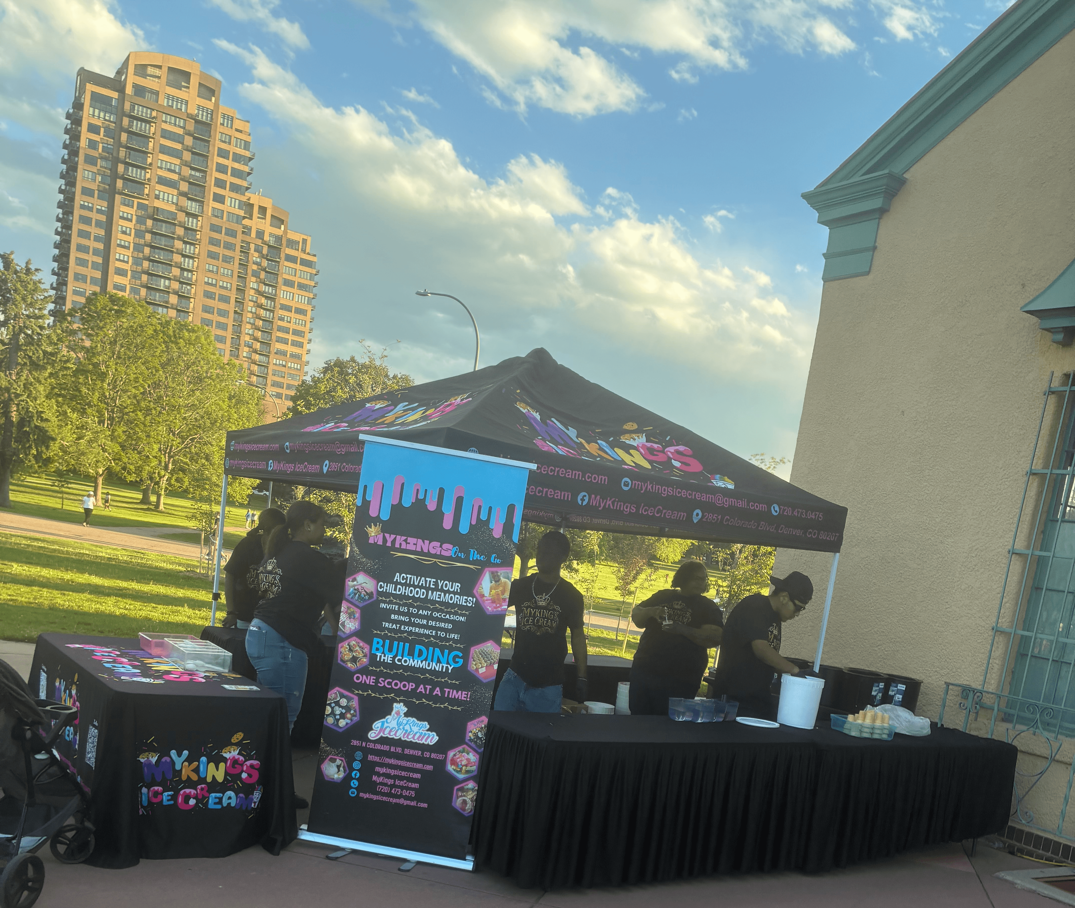 Outdoor event booth with a black canopy, people gathered around, city buildings and trees in the background, sunny sky.