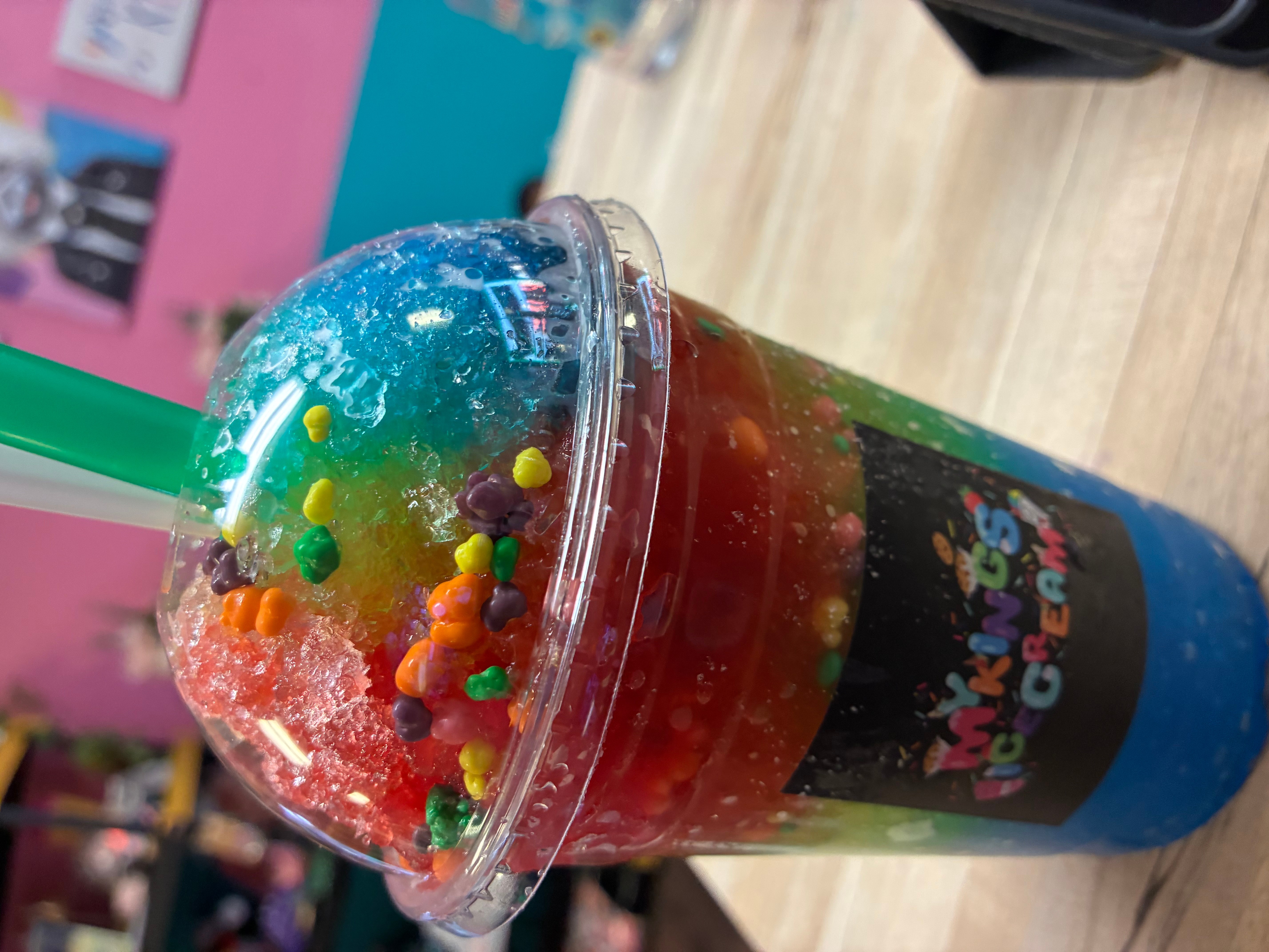 Colorful slush drink with sprinkles in a clear cup with a green straw, on a wooden surface.