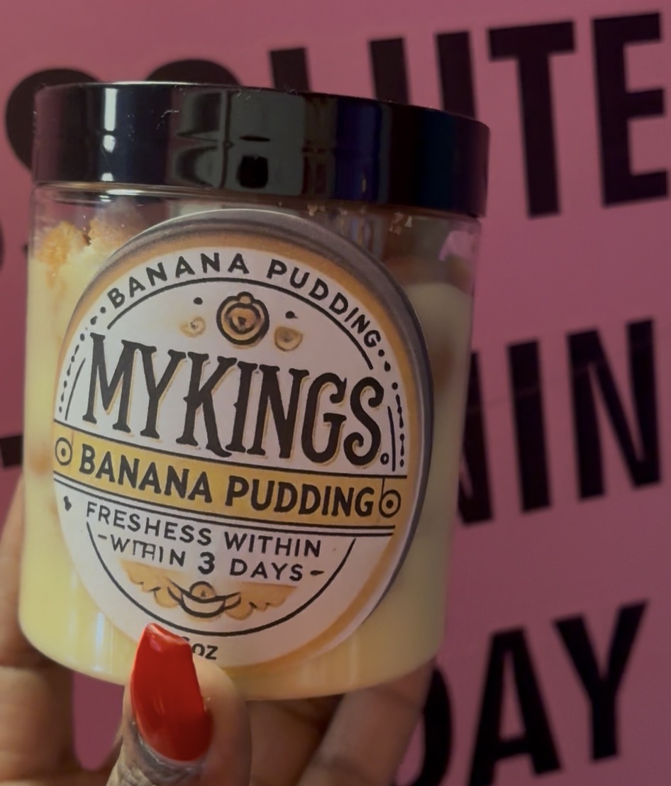 Hand holding a jar of banana pudding with a black lid, label reads MYKINGS Banana Pudding, pink background.