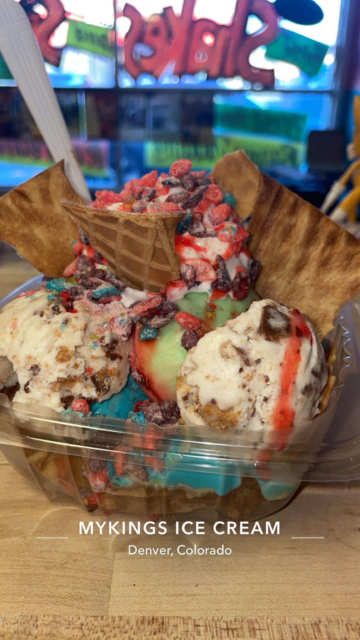 Ice cream with toppings in a clear bowl, with a waffle cone piece on top, on a wooden table.