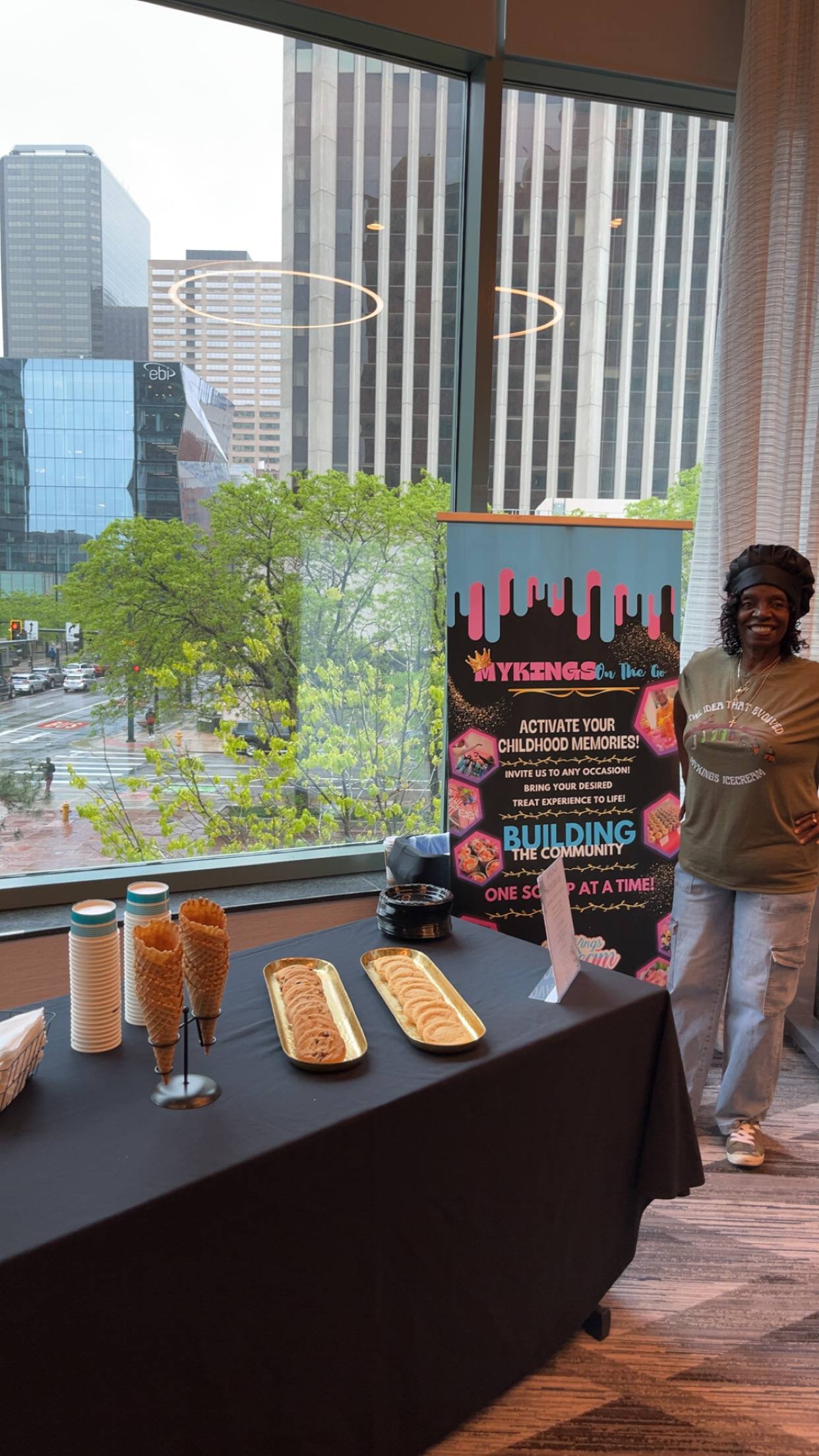 Person standing next to a promotional display with city buildings and trees outside the window, table with drinks and snacks in foreground.