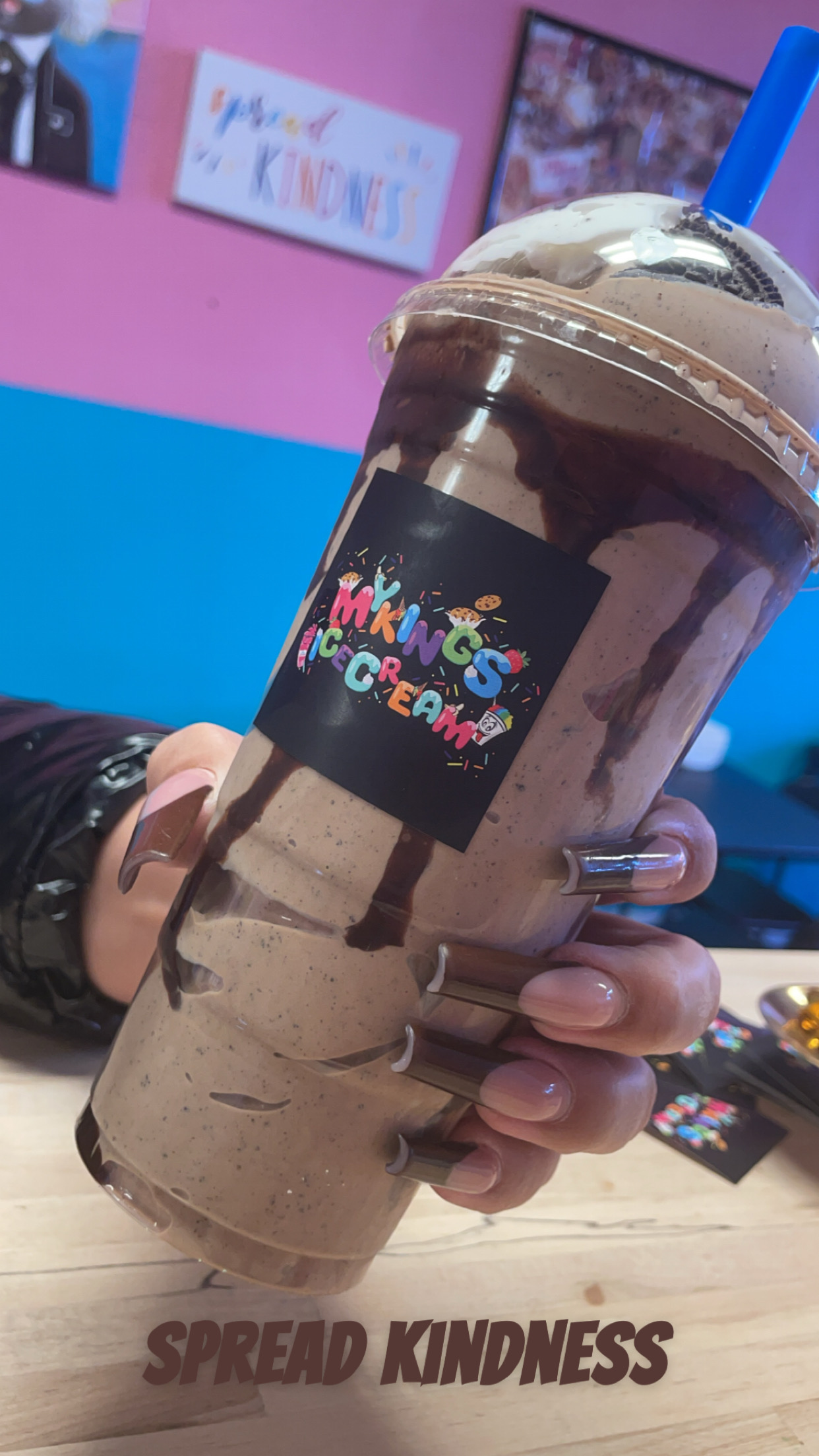 Person holding a large chocolate milkshake with a blue straw, background with pink and blue walls, and framed pictures.