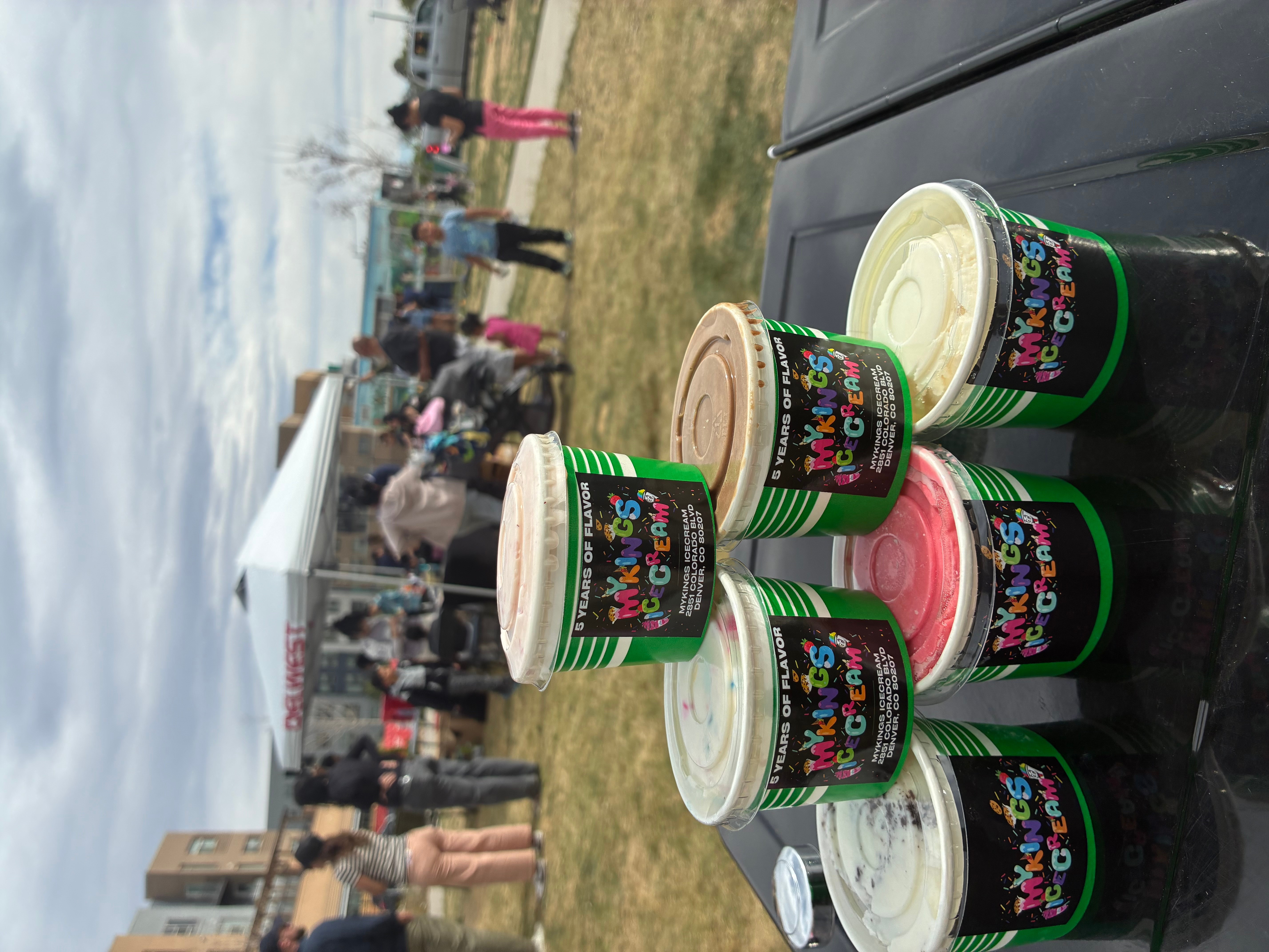 Six cups with colorful designs on a black surface, with a crowd and tents in the background outdoors.