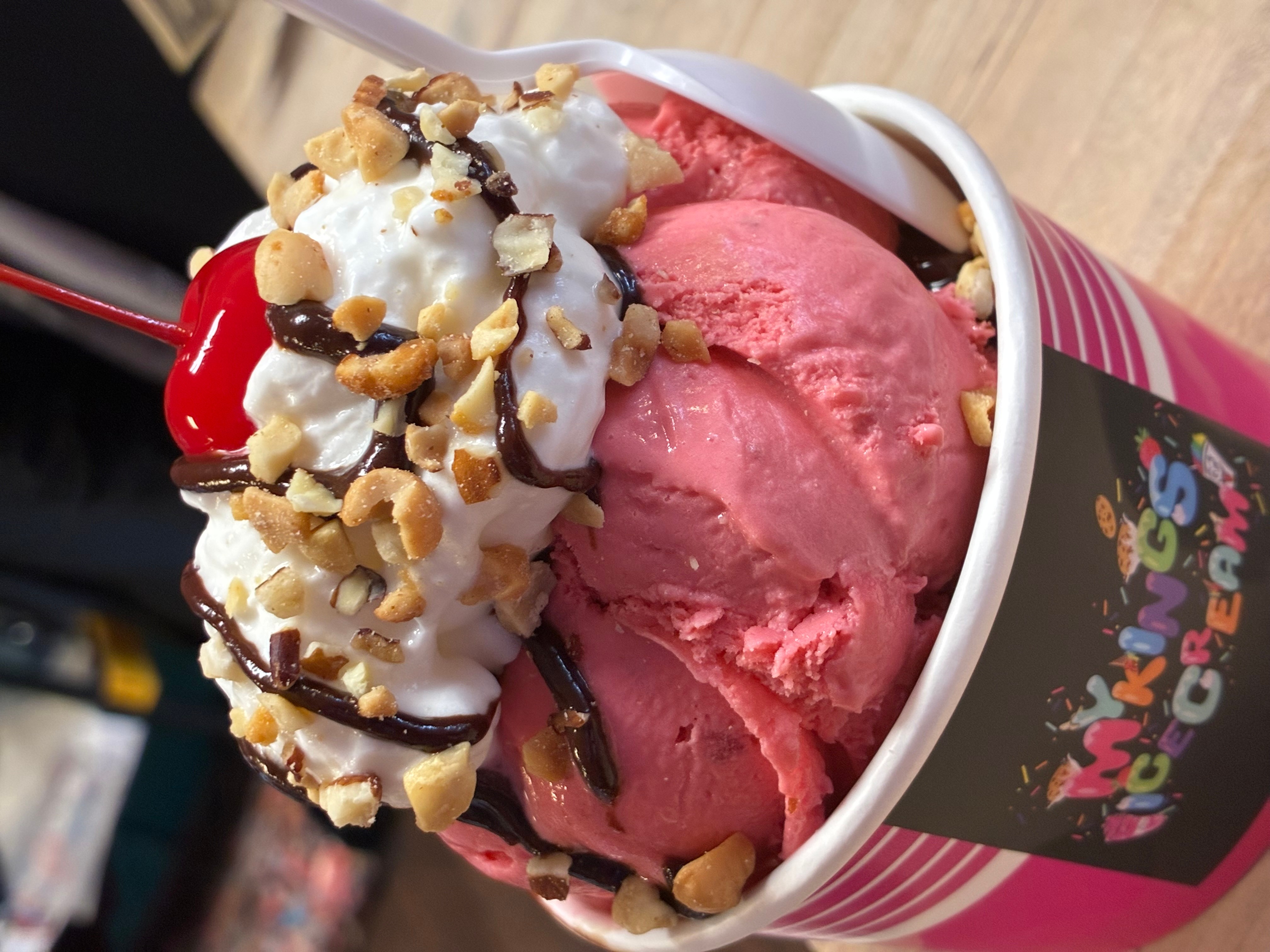 Cup of pink ice cream topped with whipped cream, nuts, and chocolate syrup, in a striped cup with colorful text.