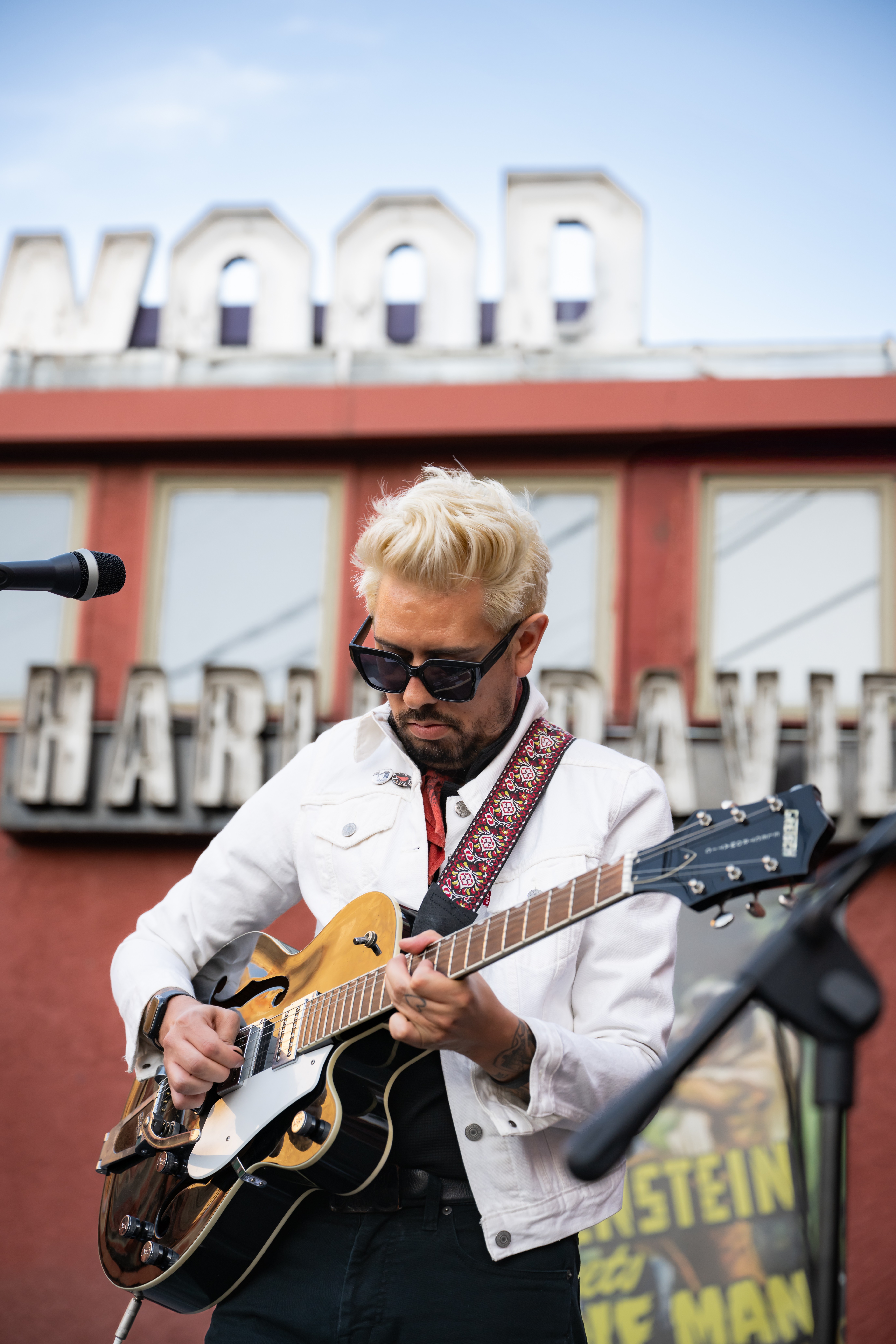 Man with blonde hair and sunglasses playing guitar outdoors, with a building and a sign in the background