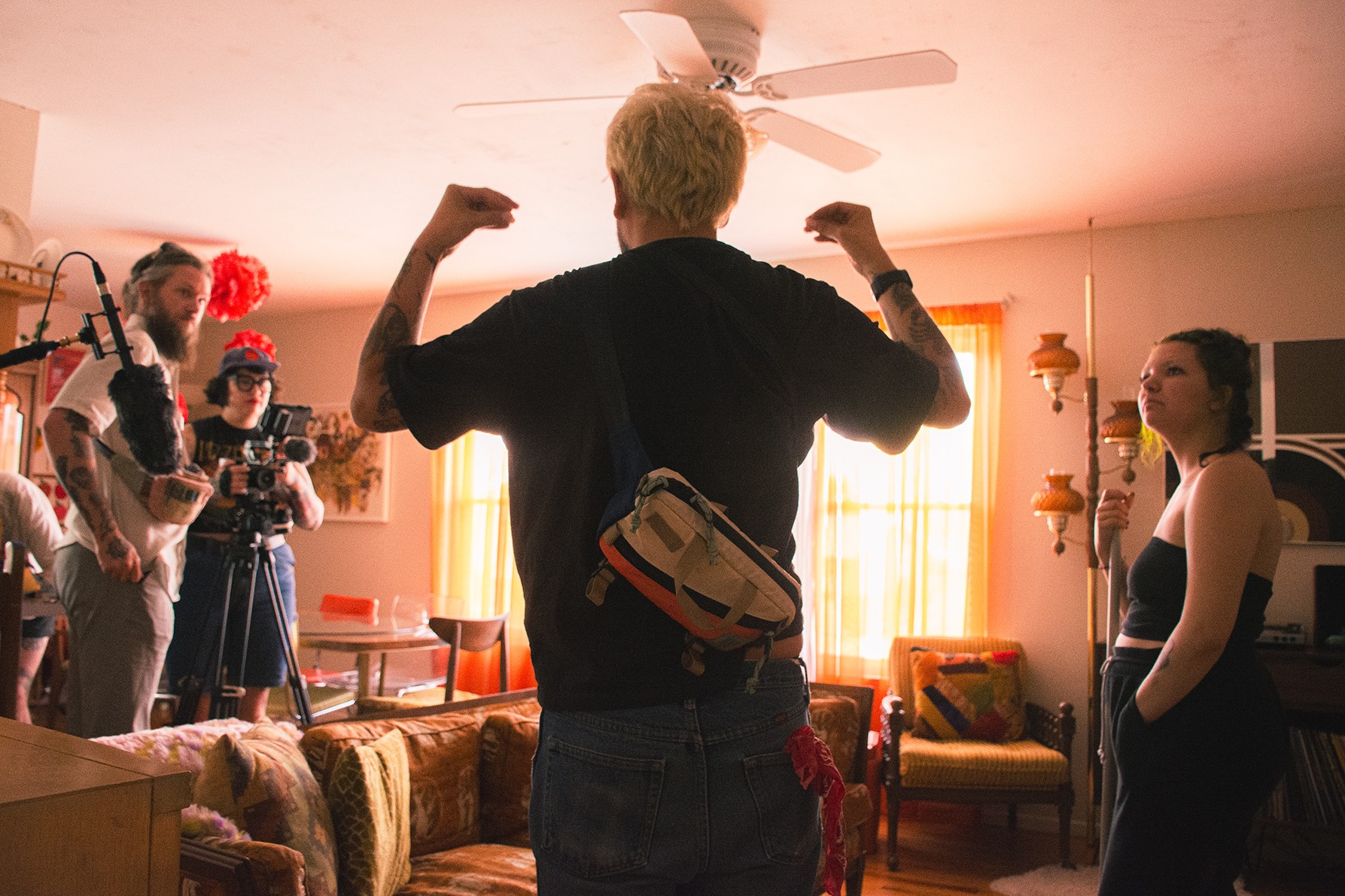Person with short hair and backpack flexing arms in a room with people and cameras, sunlight through windows, ceiling fan overhead.
