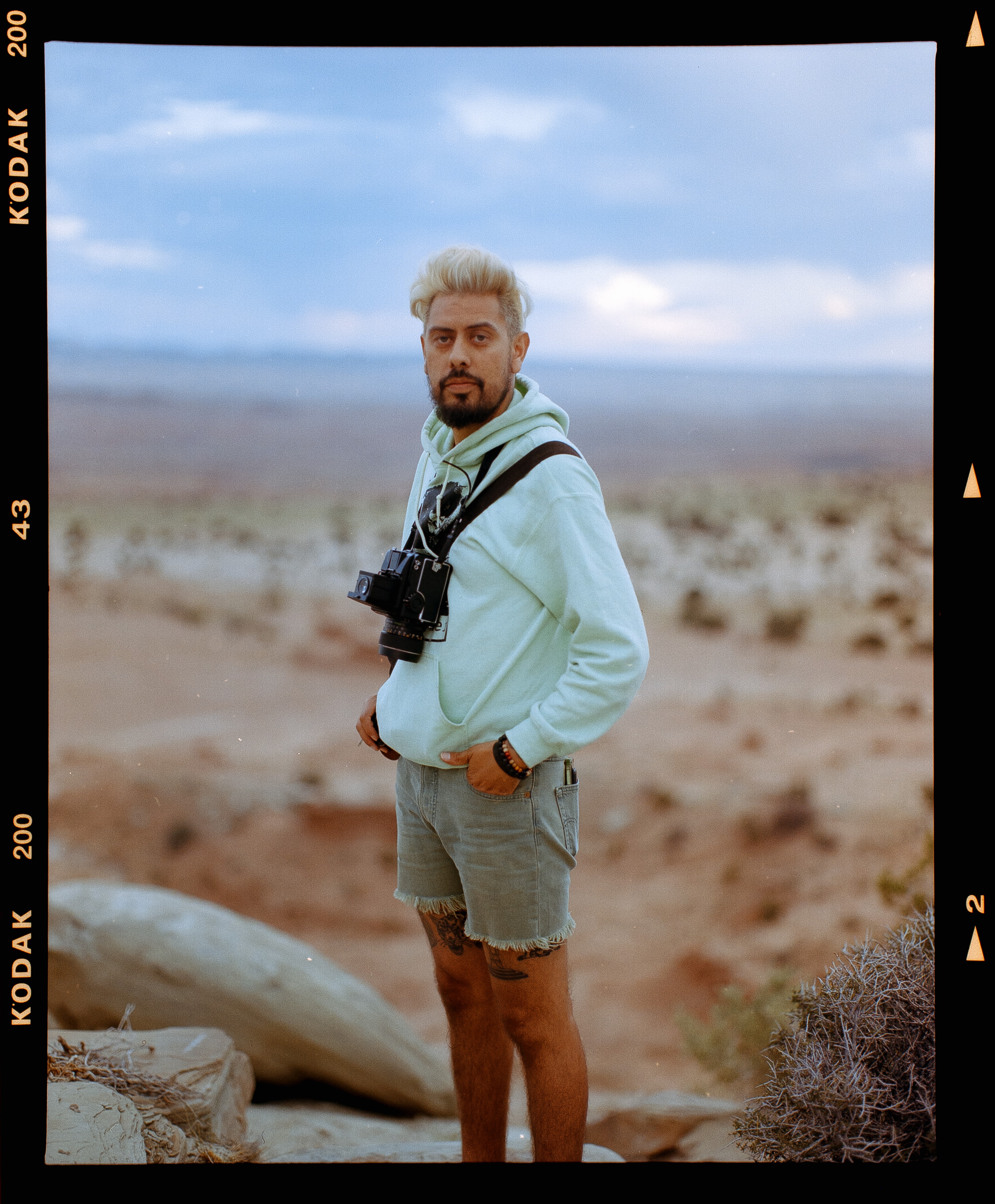 Man with gray hair and beard stands outdoors in a desert landscape, wearing a light hoodie, shorts, and holding a camera.