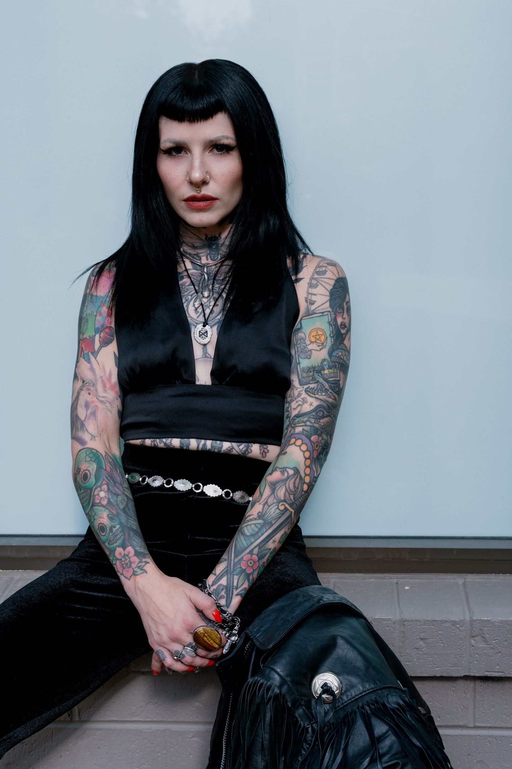 Woman with long black hair and tattoos on arms, sitting against a plain wall, wearing dark clothing and jewelry.