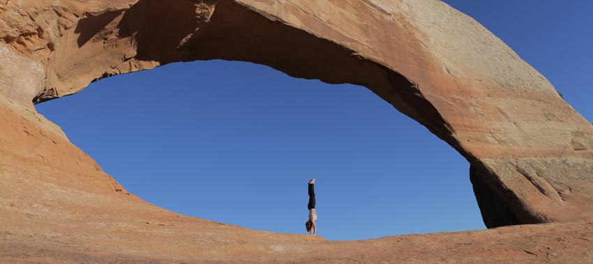 Person standing under a large natural rock arch against a clear blue sky.
