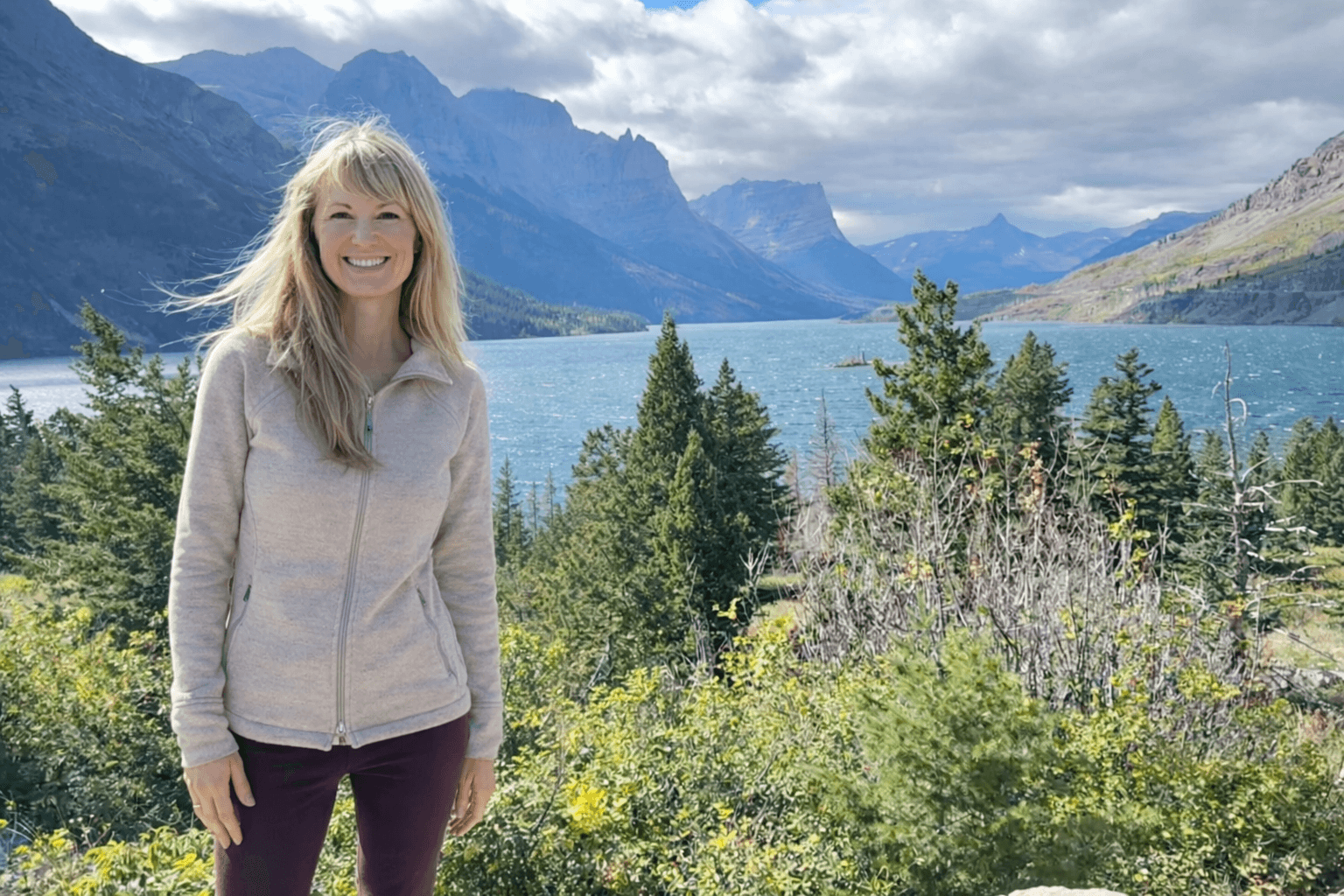 Woman standing outdoors near a lake with mountains and trees in the background, smiling, wearing a light-colored jacket.
