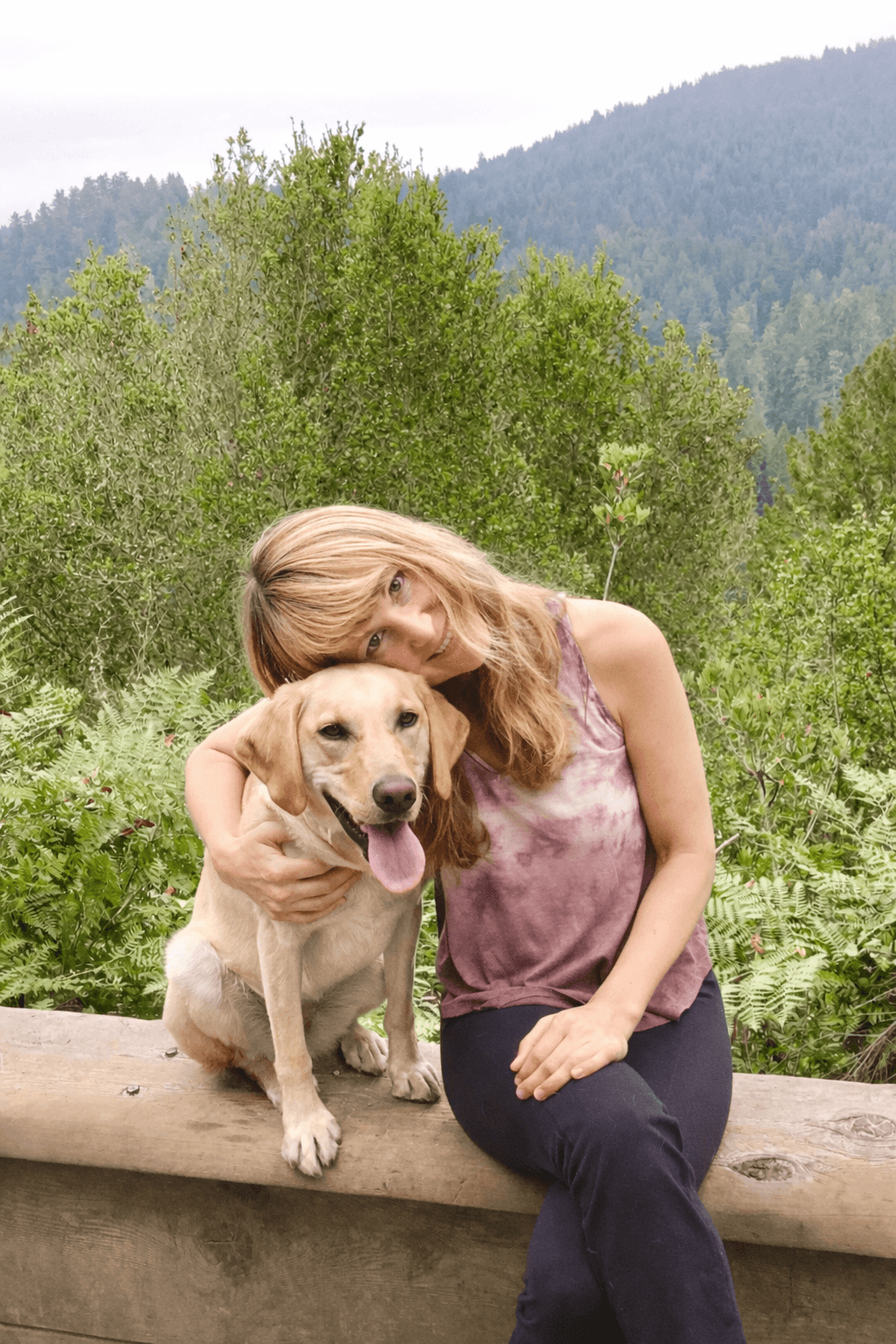 Woman with blonde hair hugging a yellow Labrador Retriever outdoors with trees and mountains in background.