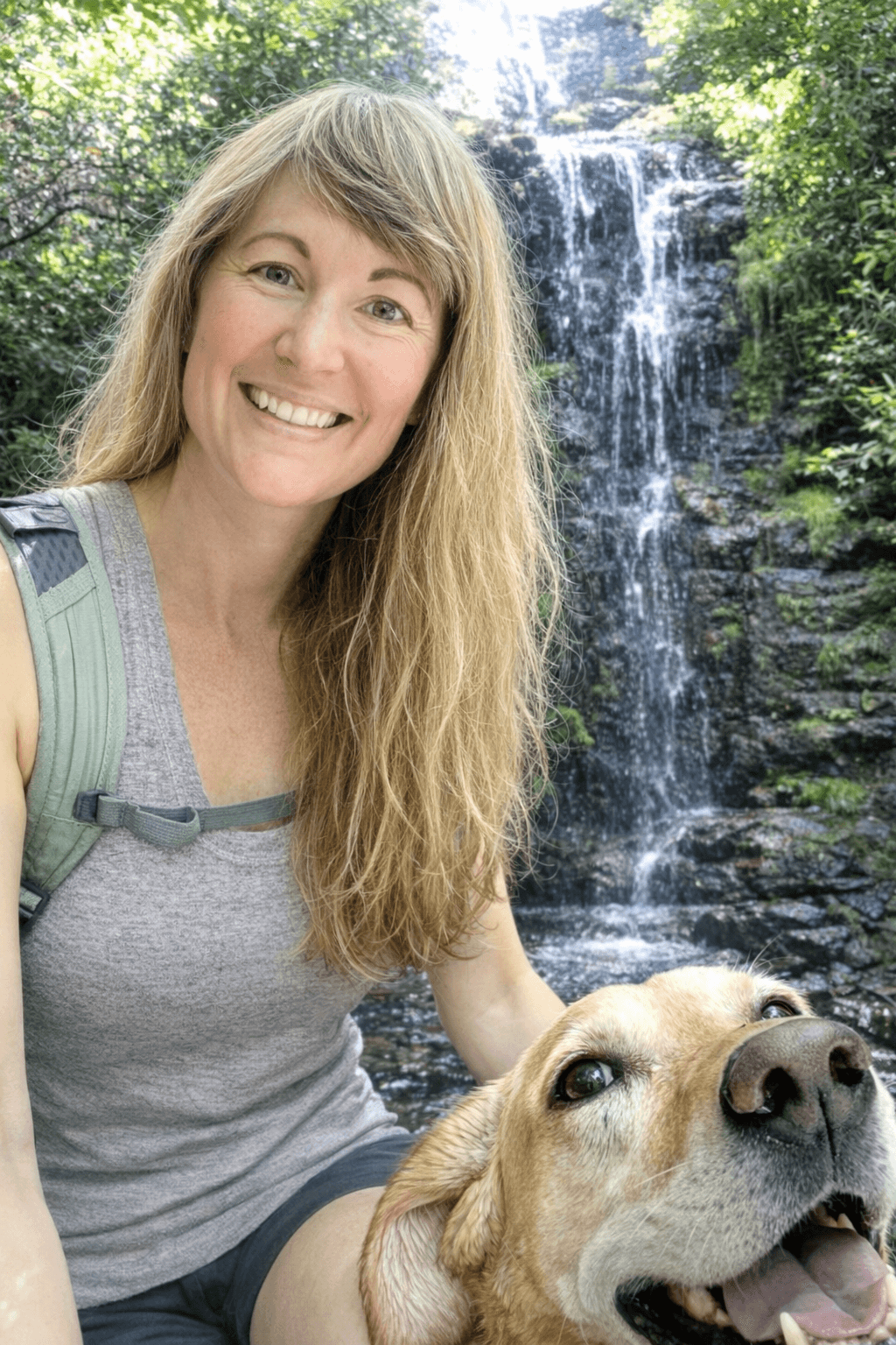 Woman with long blonde hair smiling outdoors near a waterfall with a dog, surrounded by greenery.