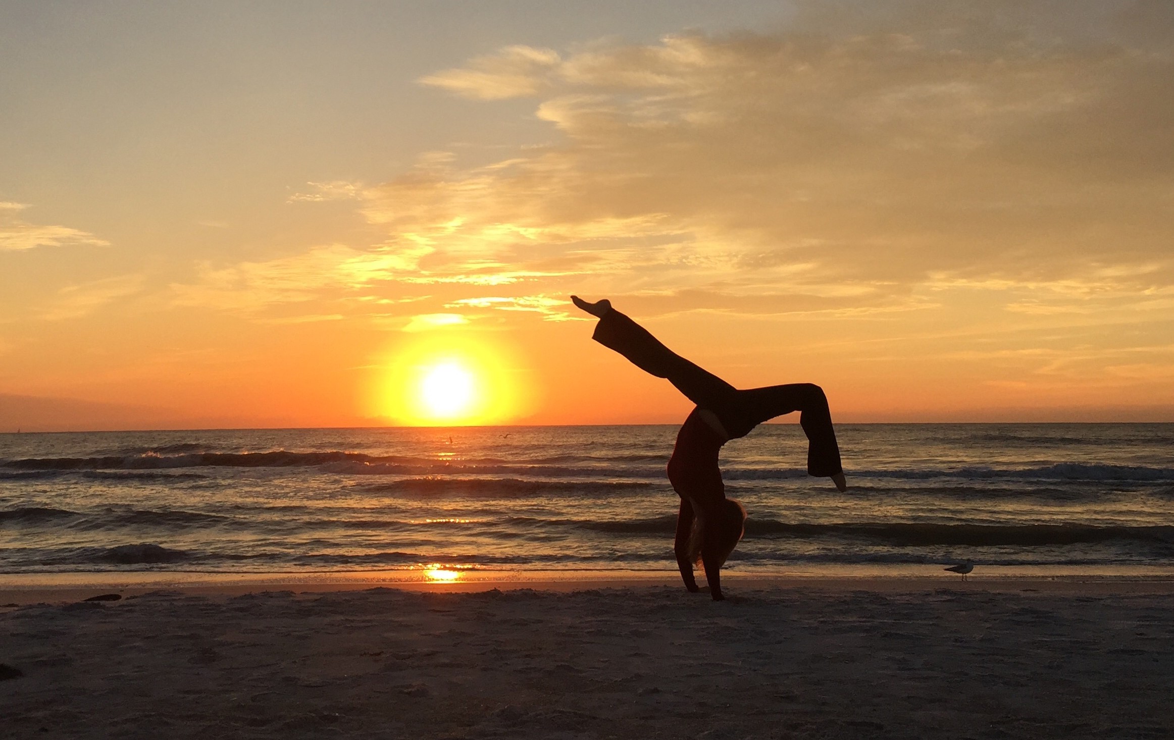 Person performing a handstand on the beach during sunset, with the sun low on the horizon and the sky partly cloudy.