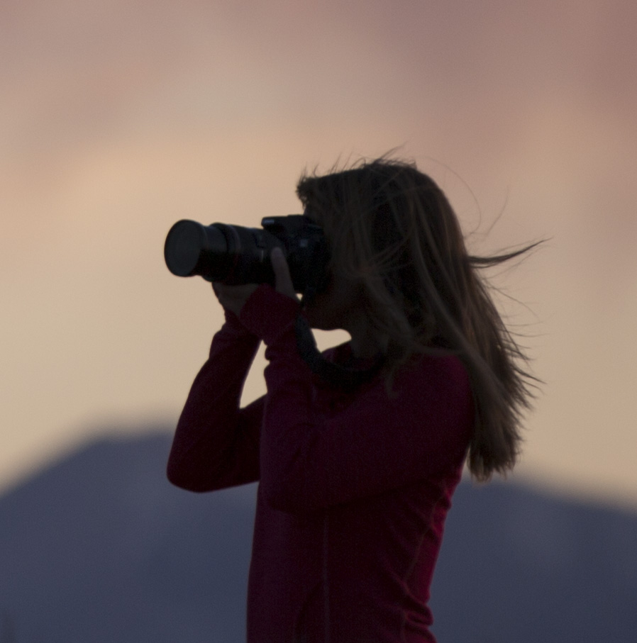 Silhouette of a person holding a camera, with a sunset sky and mountains in the background.