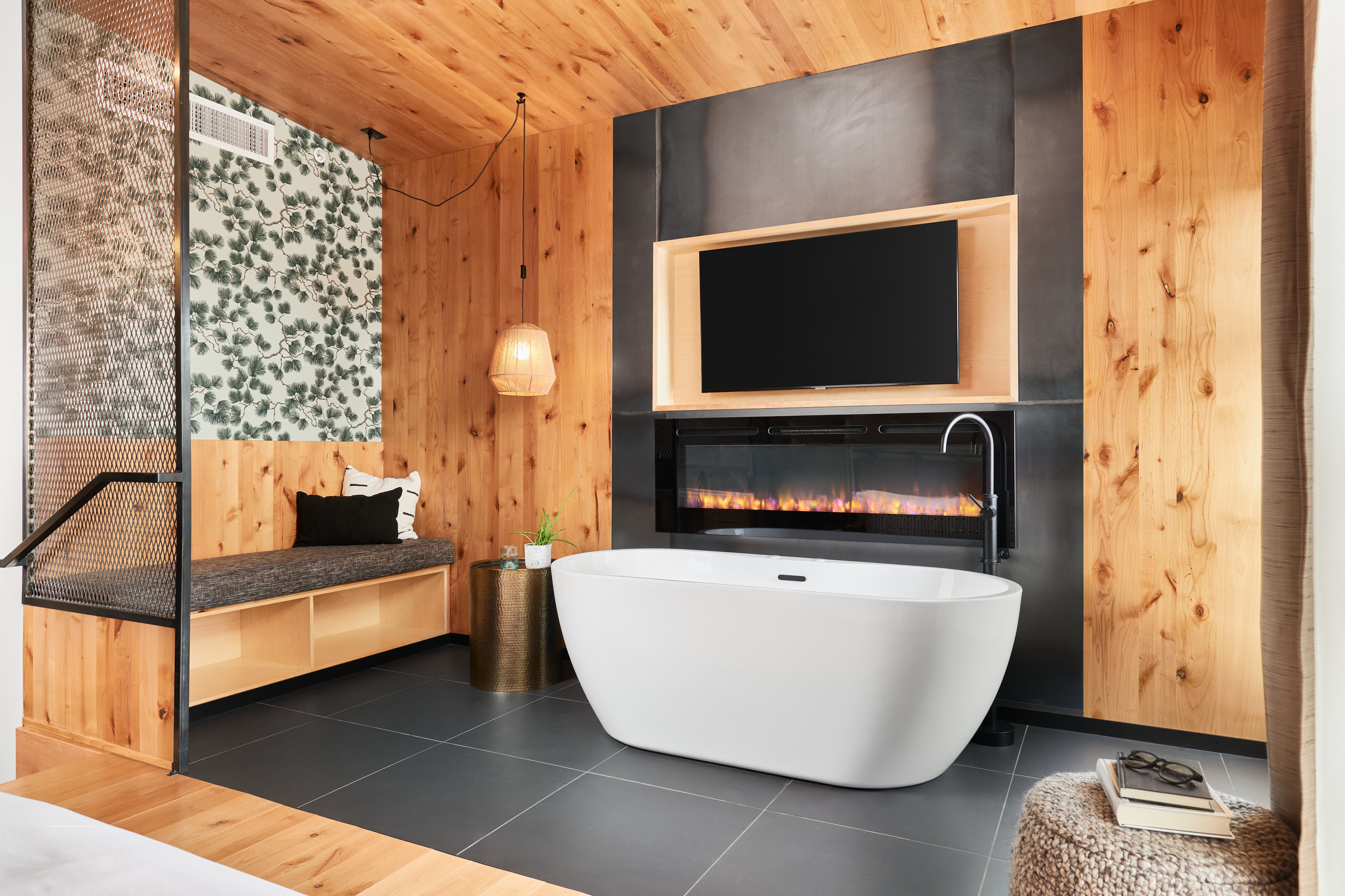 Modern bathroom with a freestanding bathtub, wooden walls, a wall-mounted TV, and a black accent wall with a fireplace.