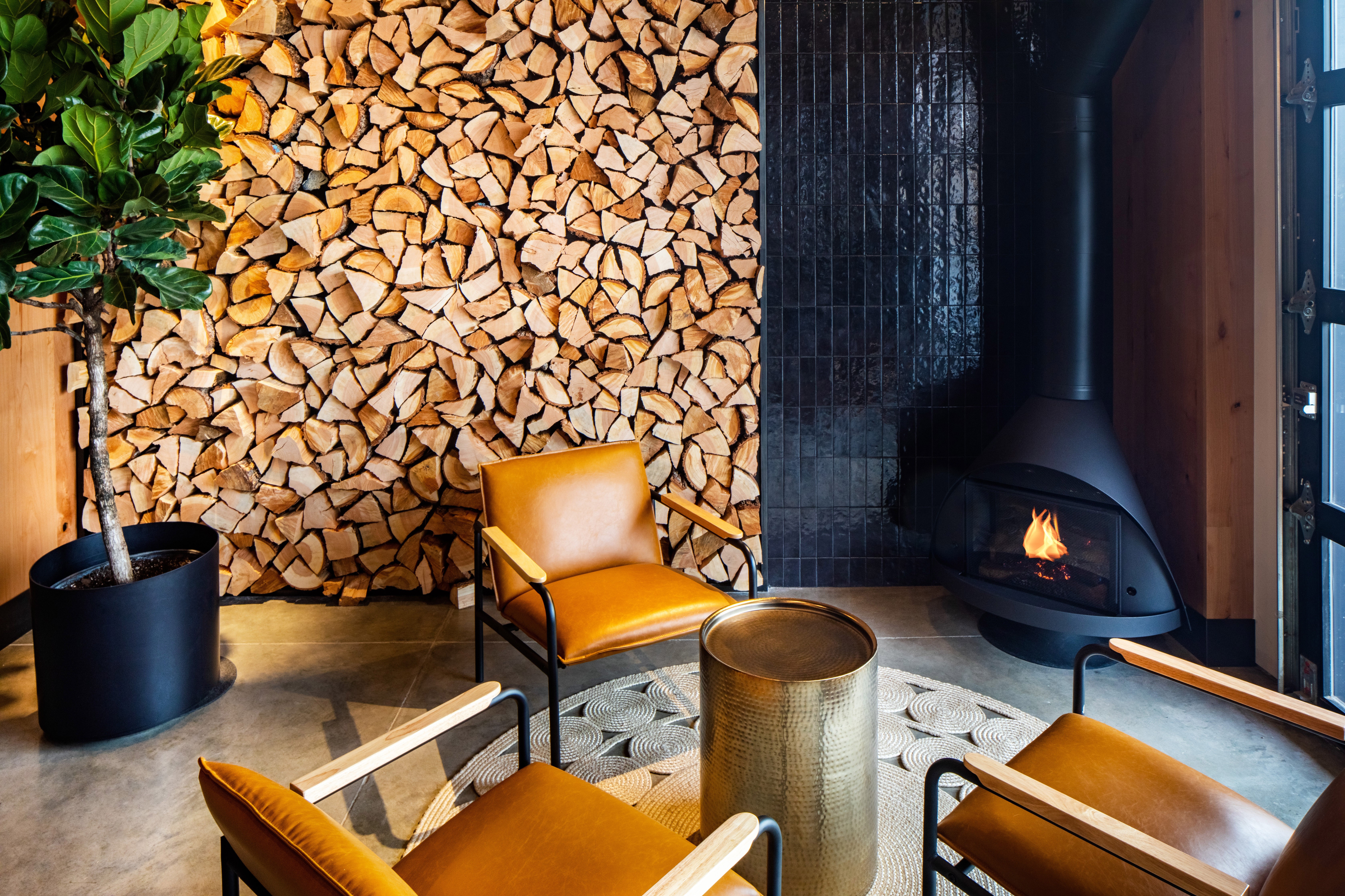 Interior with stacked firewood wall, yellow armchairs, a potted plant, and a black fireplace with a fire burning.
