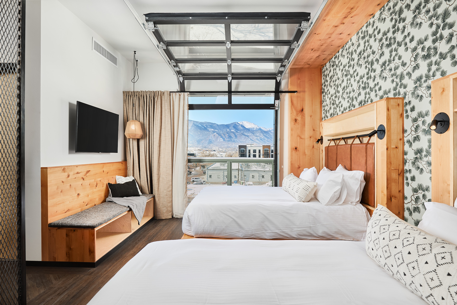 Two beds in a modern hotel room with a large window showing mountains, wooden accents, and patterned wall, daytime.