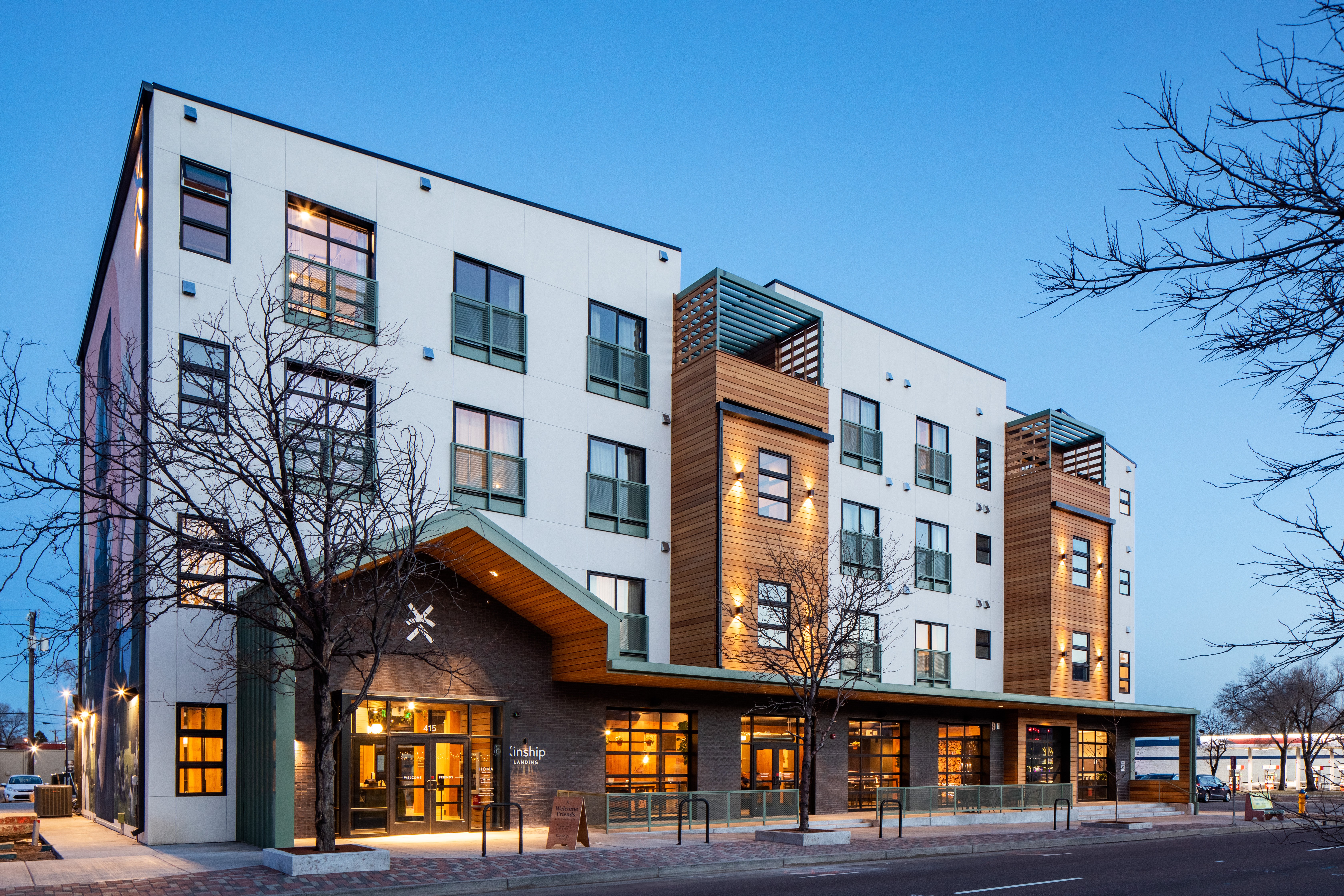 Modern multi-story building with large windows and exterior lighting, set against a clear evening sky, with trees nearby.