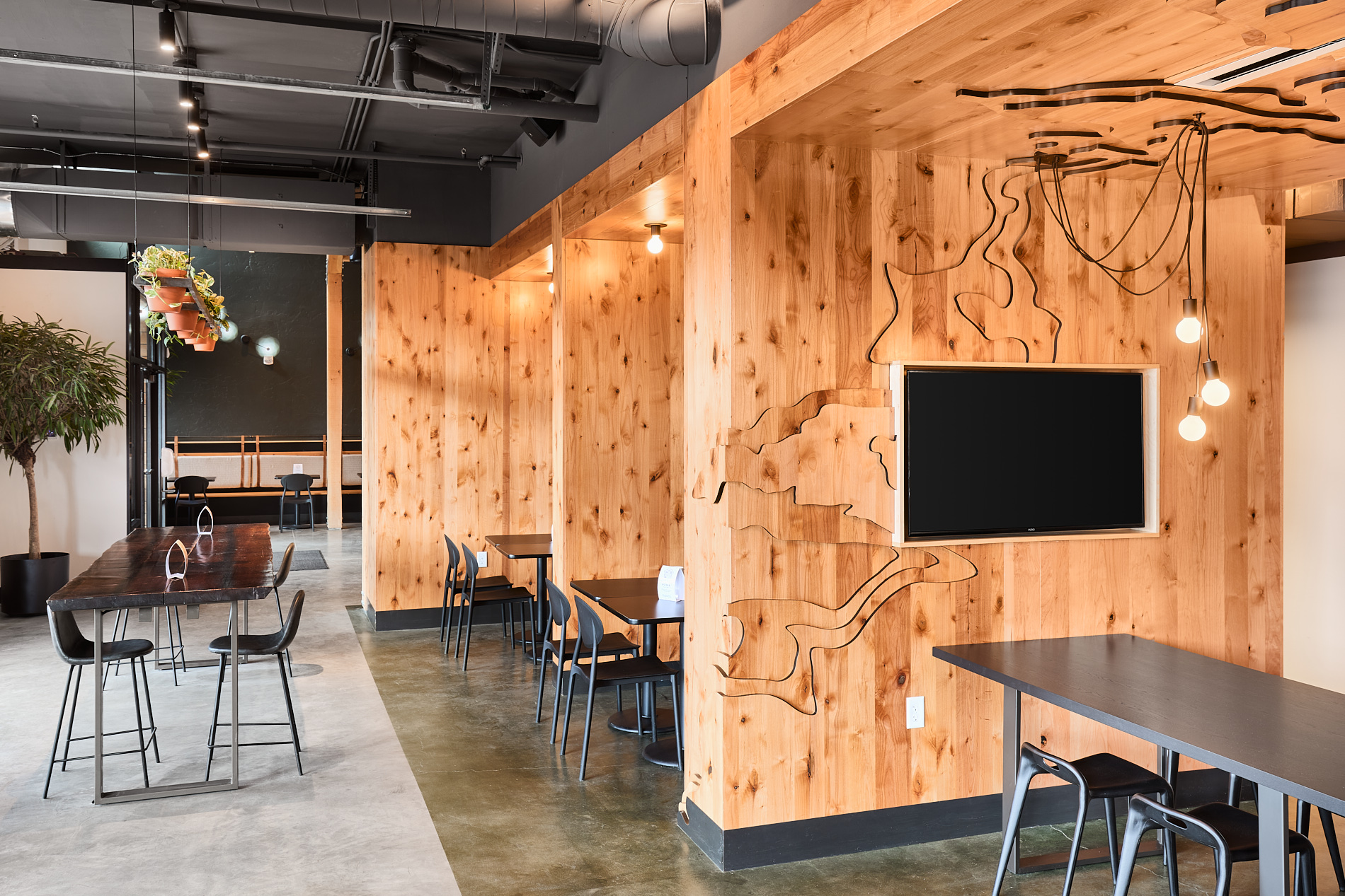 Interior of a modern cafe with wooden walls, tables, chairs, and a wall-mounted TV, with lighting and decor elements.