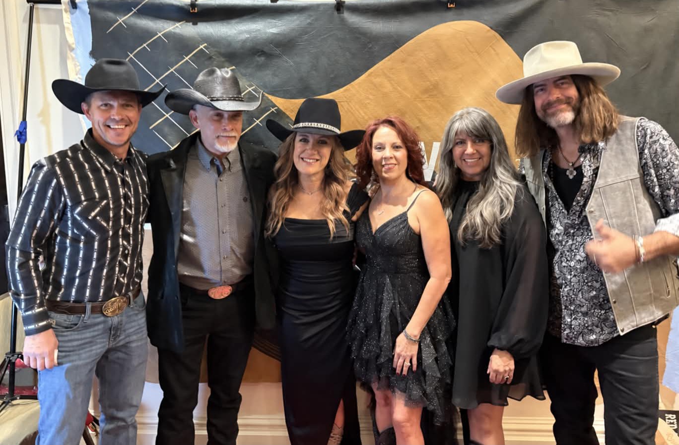 Six people standing together, smiling, in front of a black backdrop with a guitar silhouette, dressed in western-style clothing.