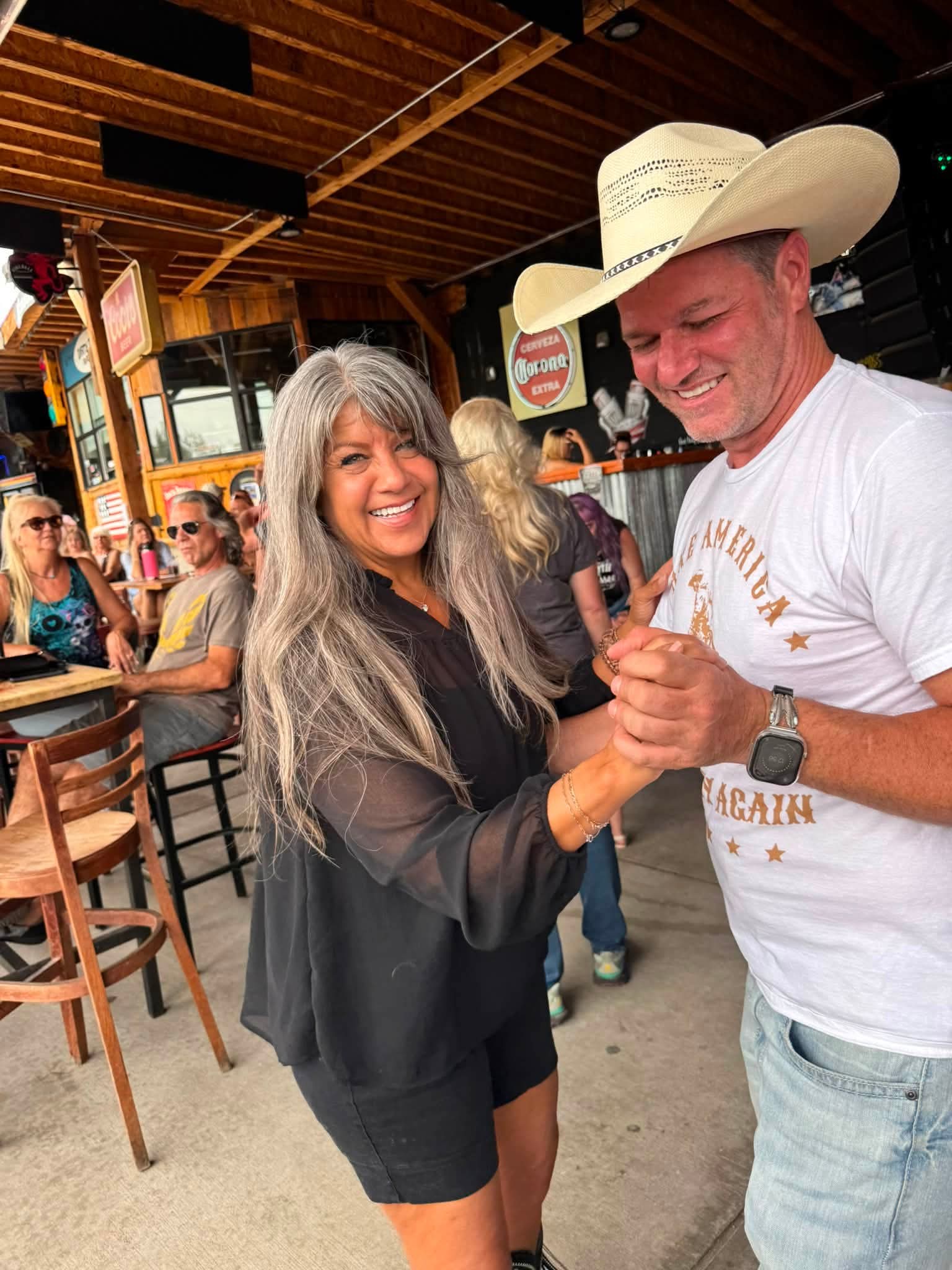A woman with long gray hair and a man wearing a cowboy hat dance together outdoors, smiling, with a crowd in the background.