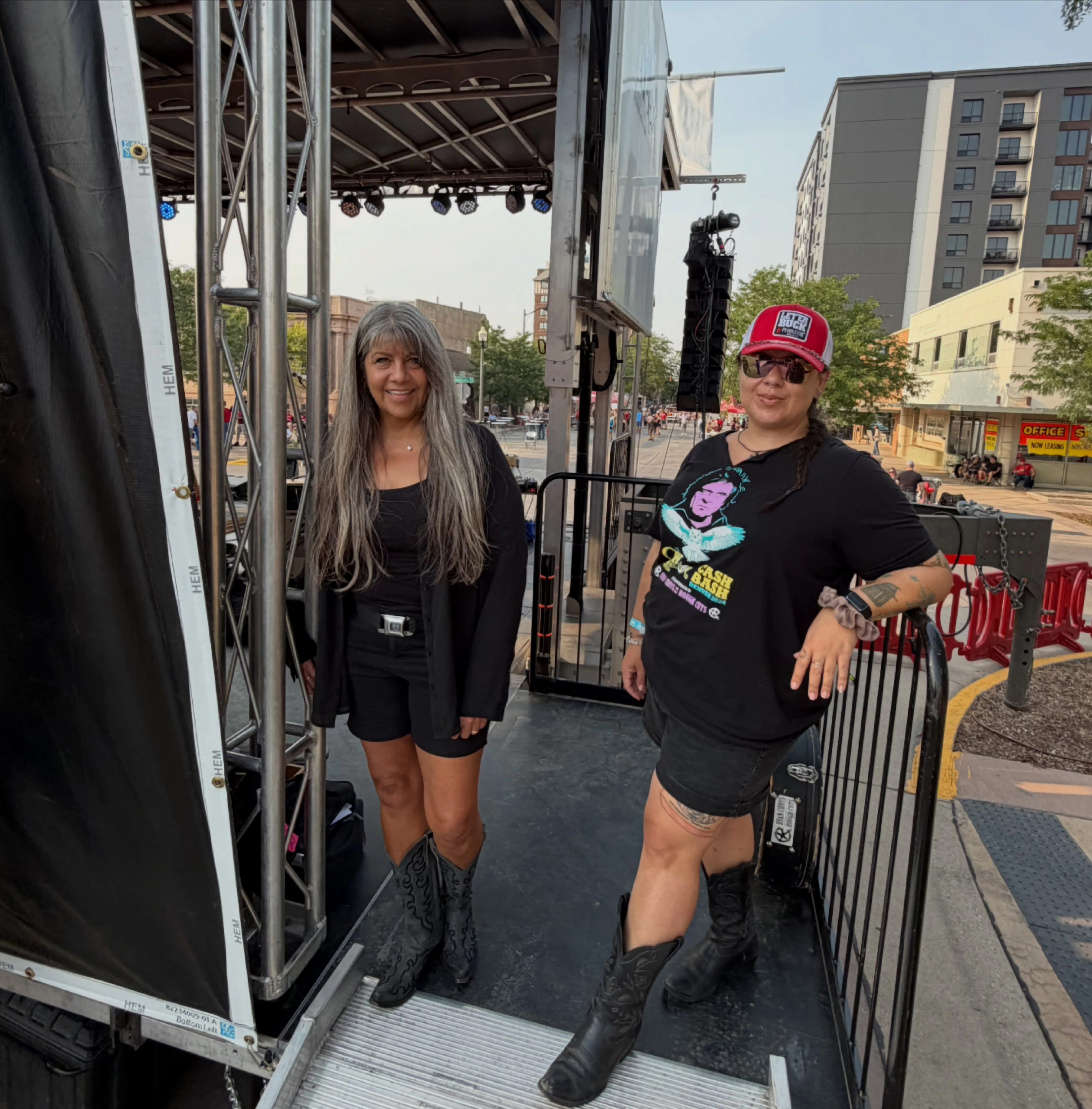 Two women standing on a stage outdoors, one with long gray hair and the other wearing a red cap and sunglasses, both smiling.