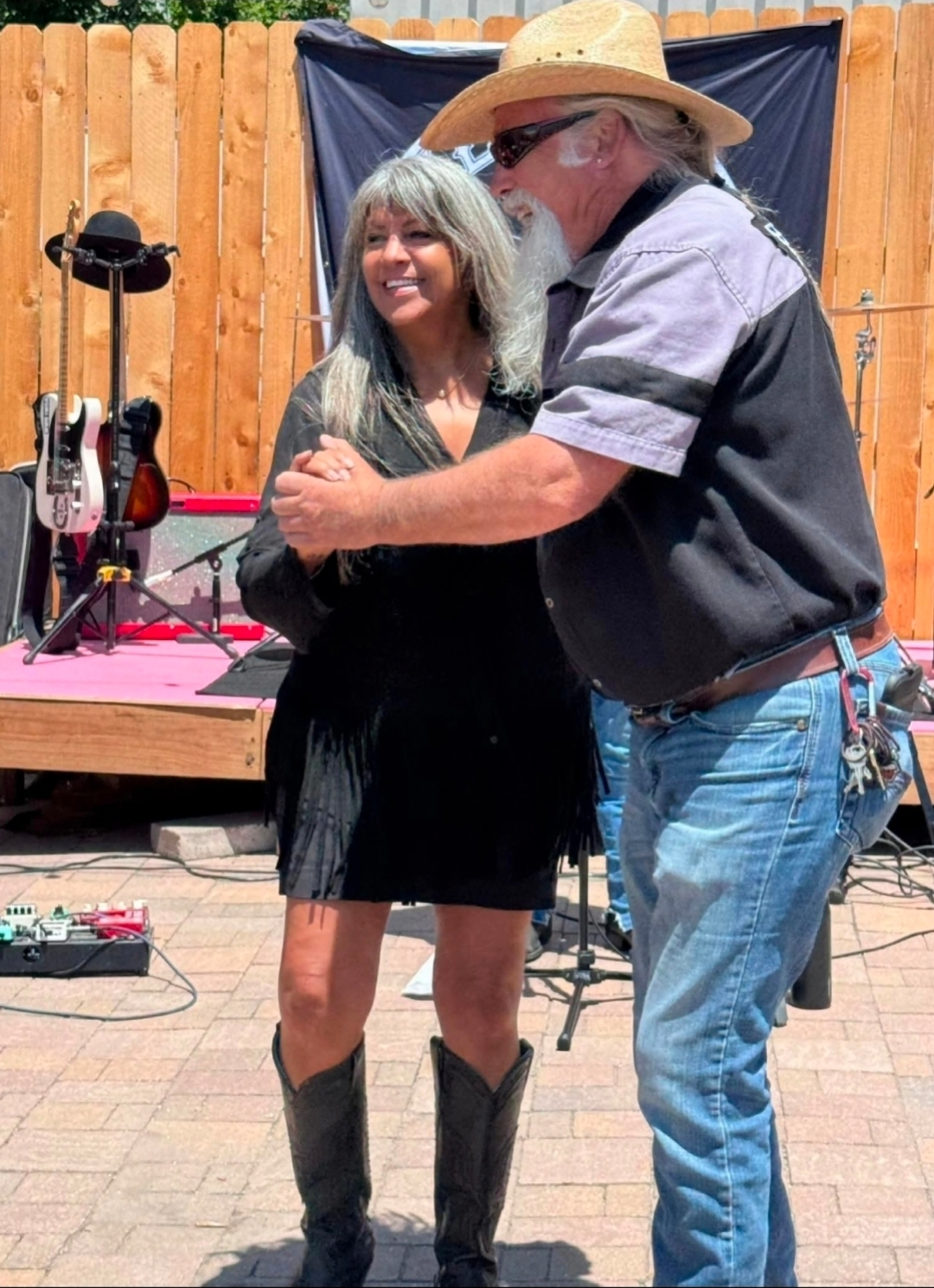A woman and a man are dancing outdoors in front of a wooden fence, with musical equipment nearby.