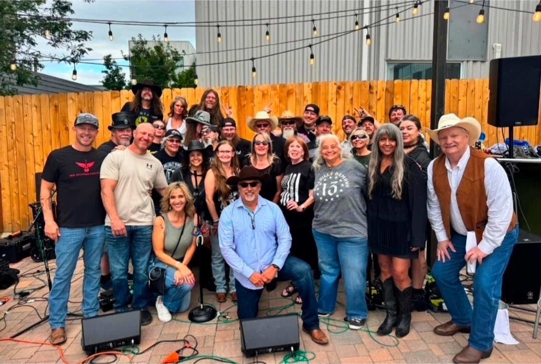 Group of people posing outdoors with string lights and a wooden fence, some wearing hats and casual clothing.