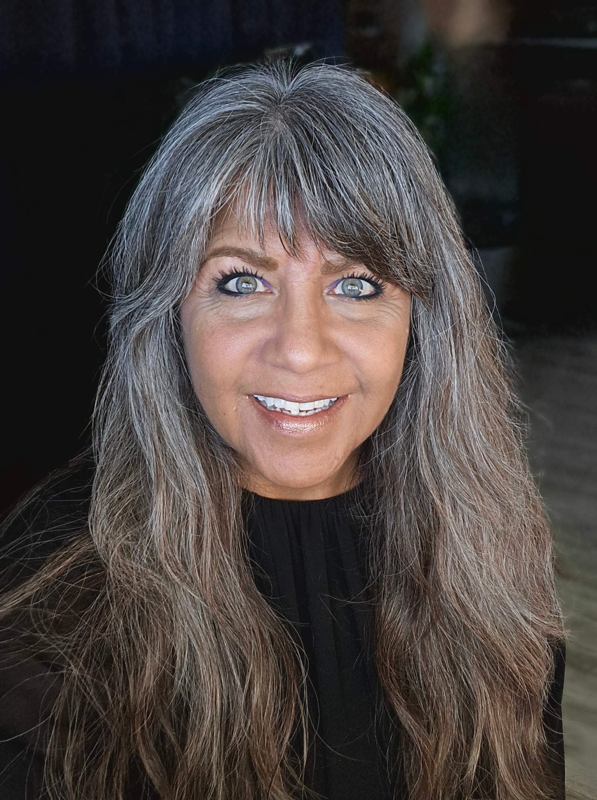 Woman with long, wavy gray hair and blue eyes smiling, wearing a black top, indoors.