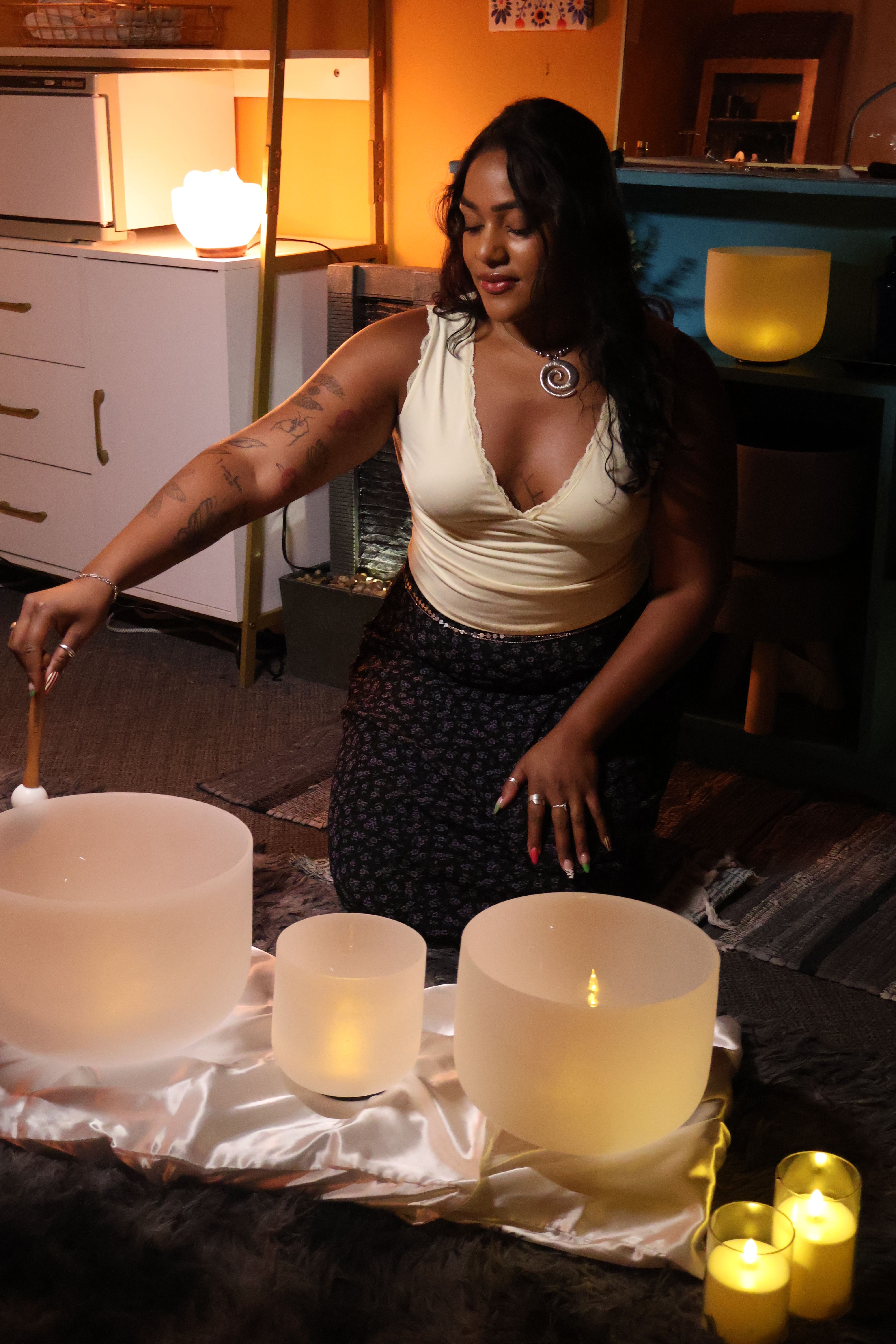 Woman with dark hair and tattoos, wearing a white top and dark skirt, standing indoors near candles on a table.