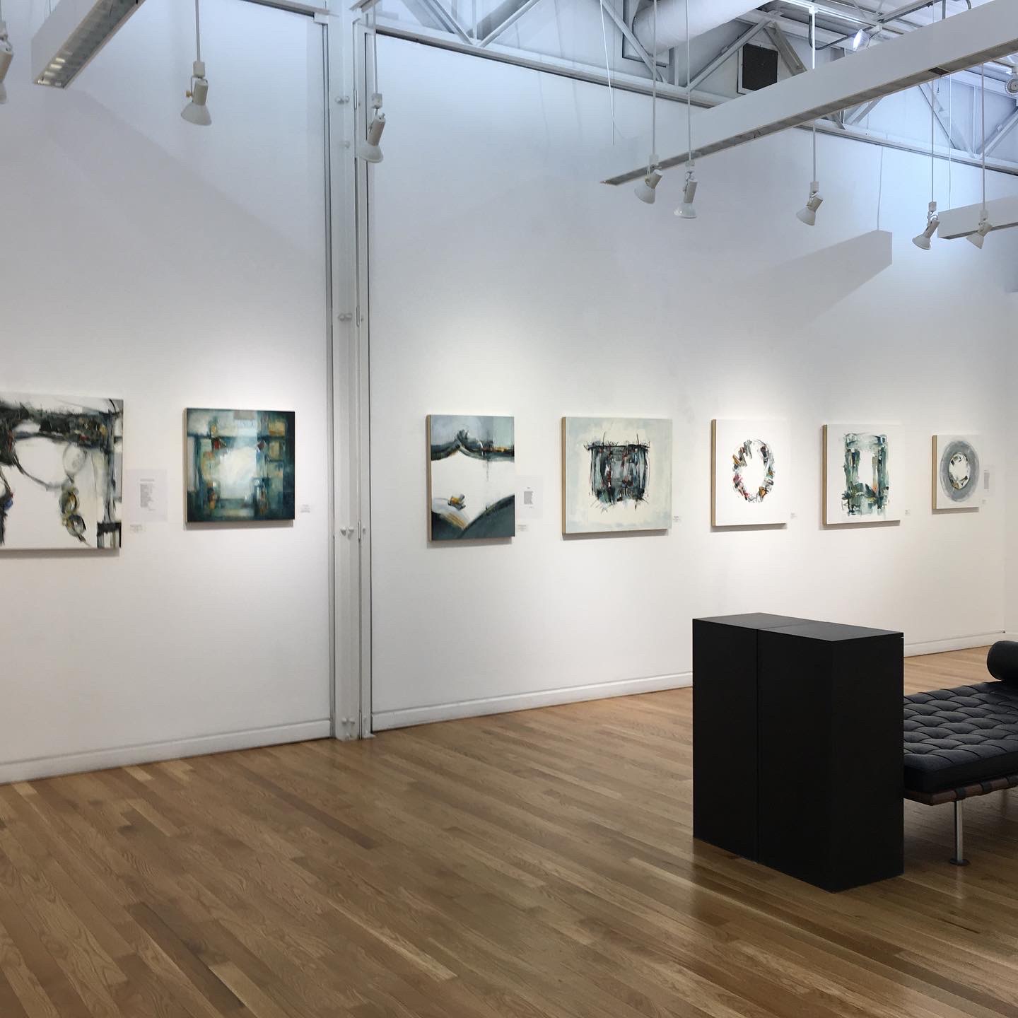 Gallery wall with abstract paintings, wooden floor, black bench, and white walls in an art gallery.