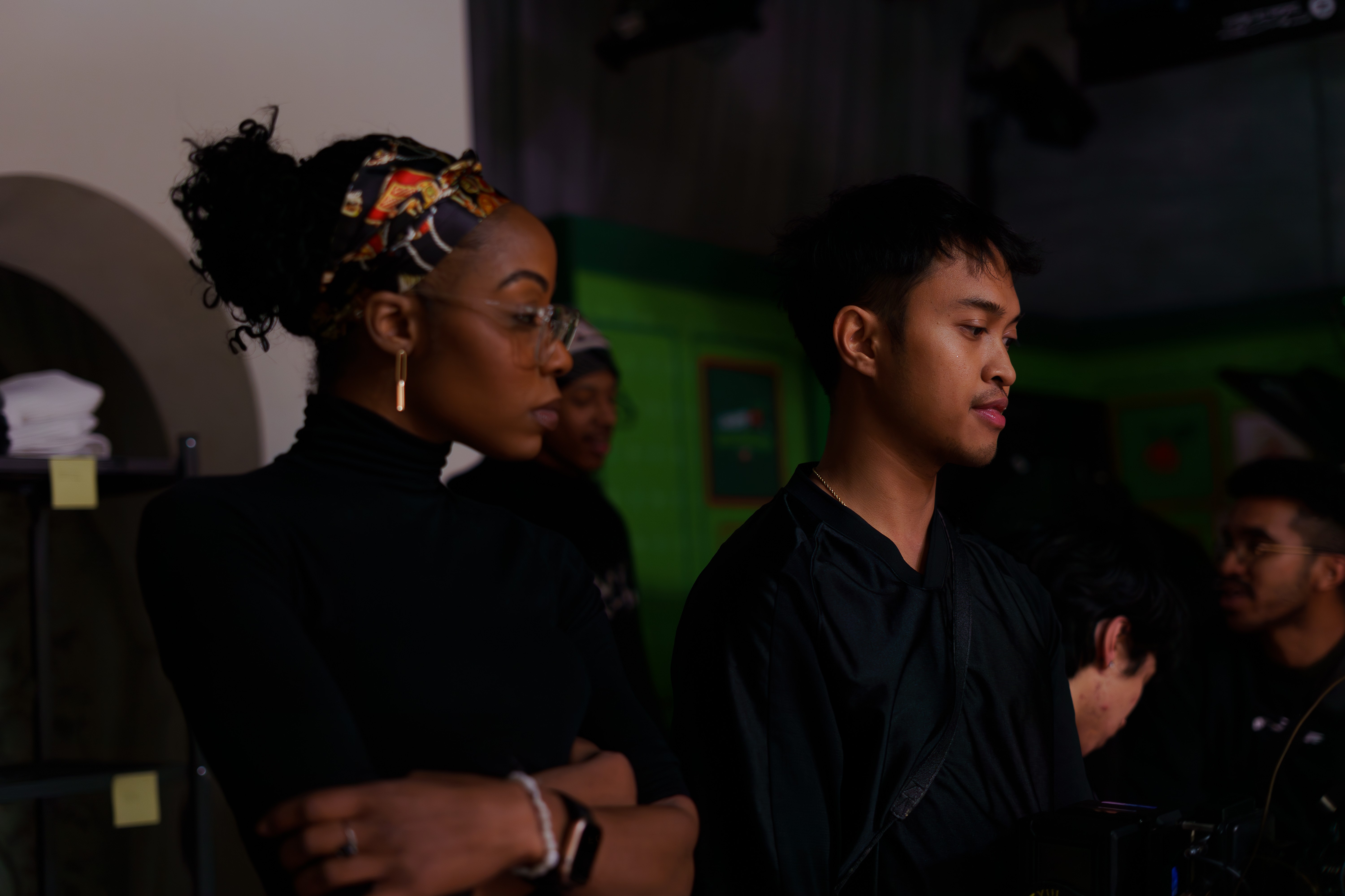 Two people standing indoors, one with glasses and a headband, the other with short hair, both looking to the right.