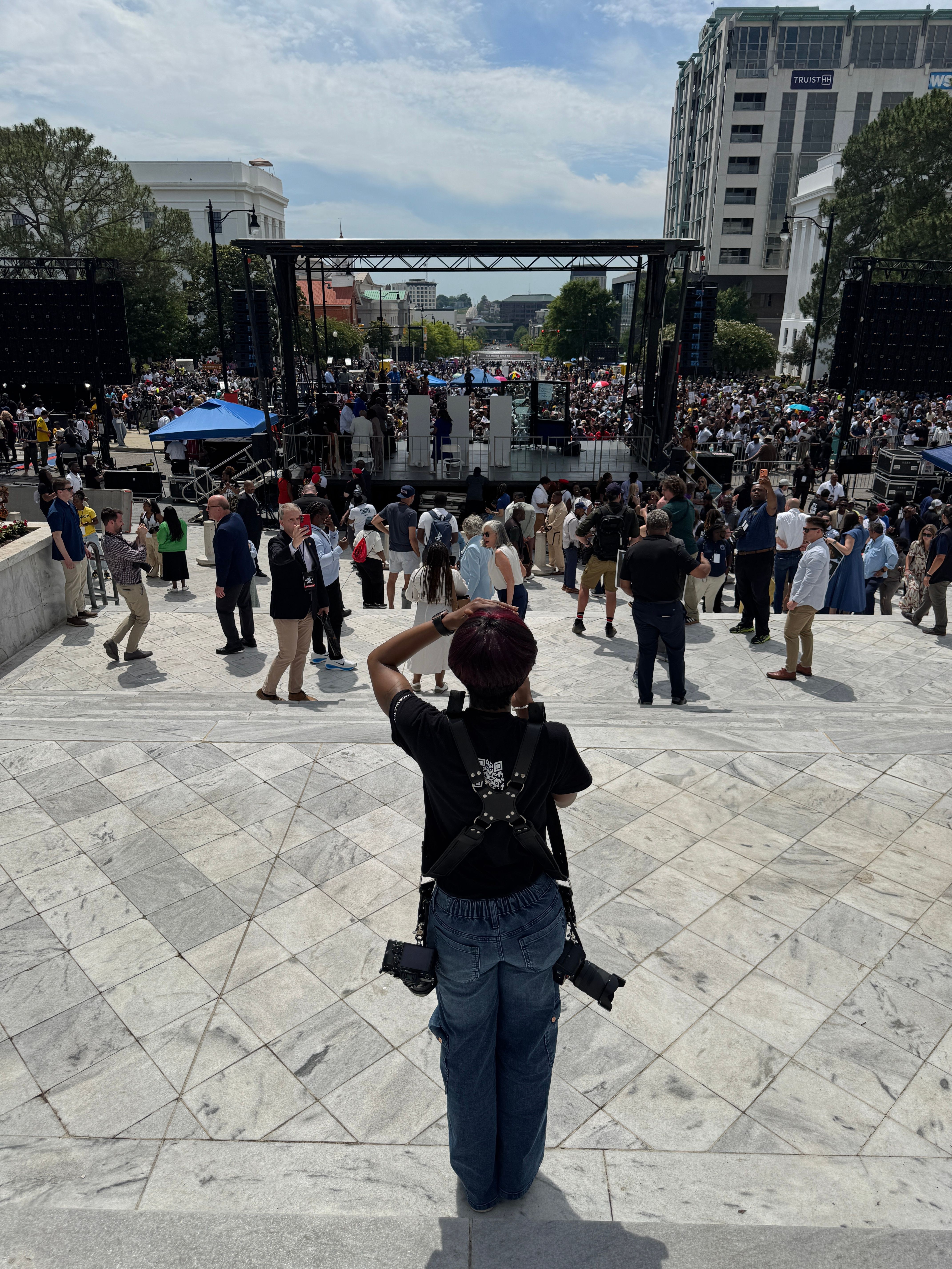 Crowd of people gathered outdoors on a paved area with buildings and trees in the background.