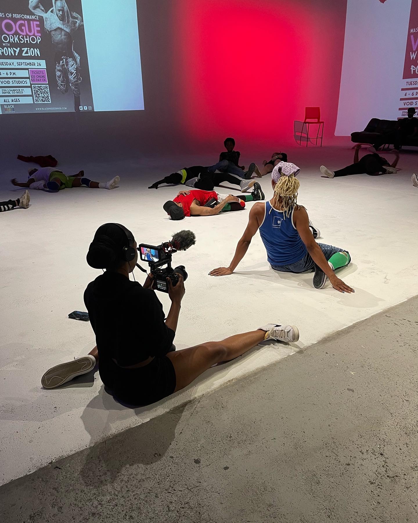 A woman with a camera sits on the floor watching a group of people lying down during a performance or workshop, with a red-lit backdrop.