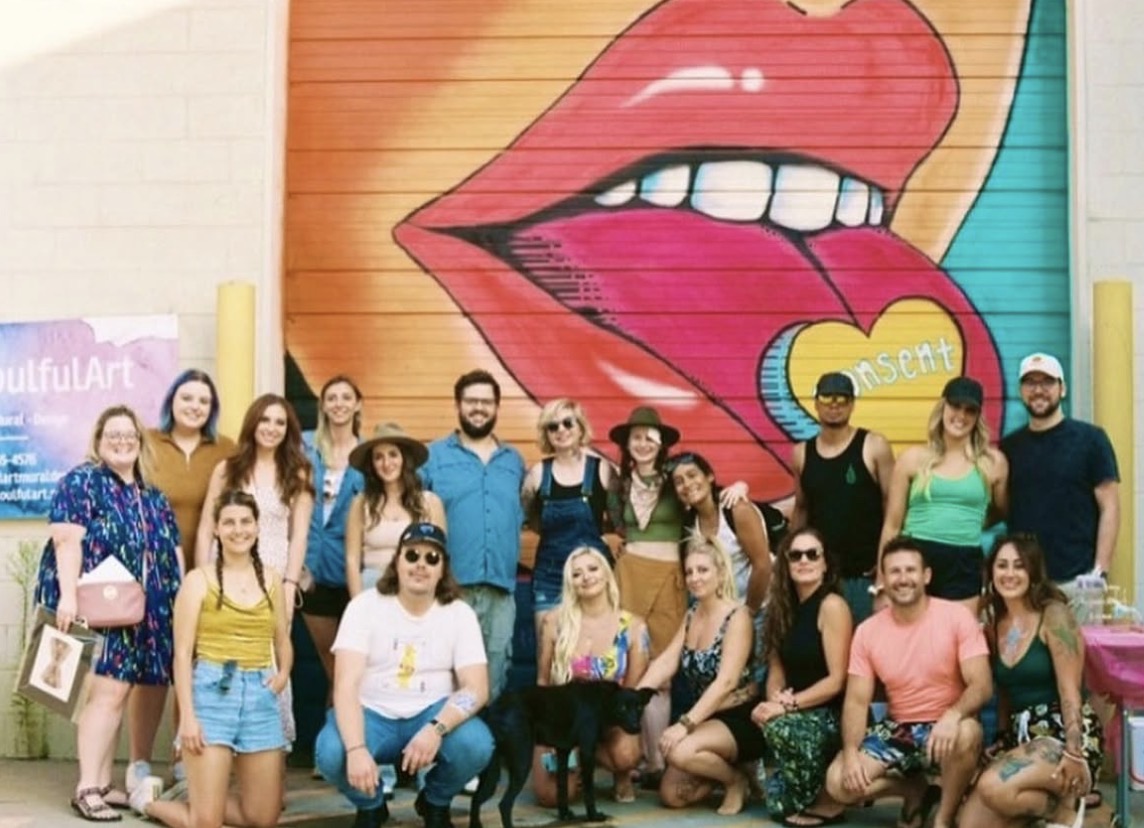 Group of people posing in front of a colorful mural with lips and a heart, some sitting and some standing.