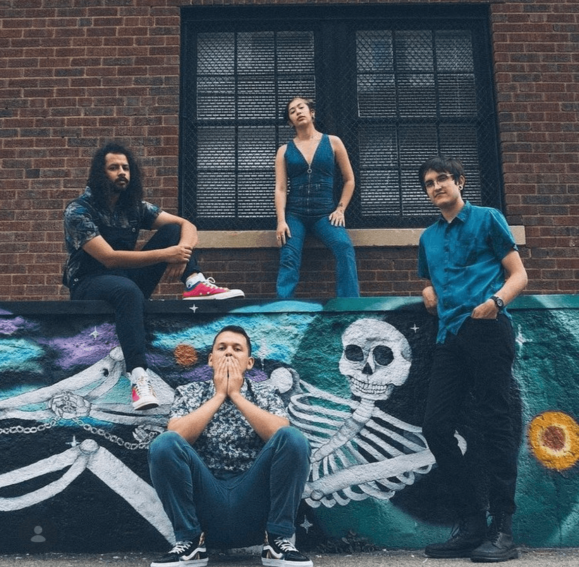 Four people pose in front of a colorful mural with a skeleton, in front of a brick building with a window.