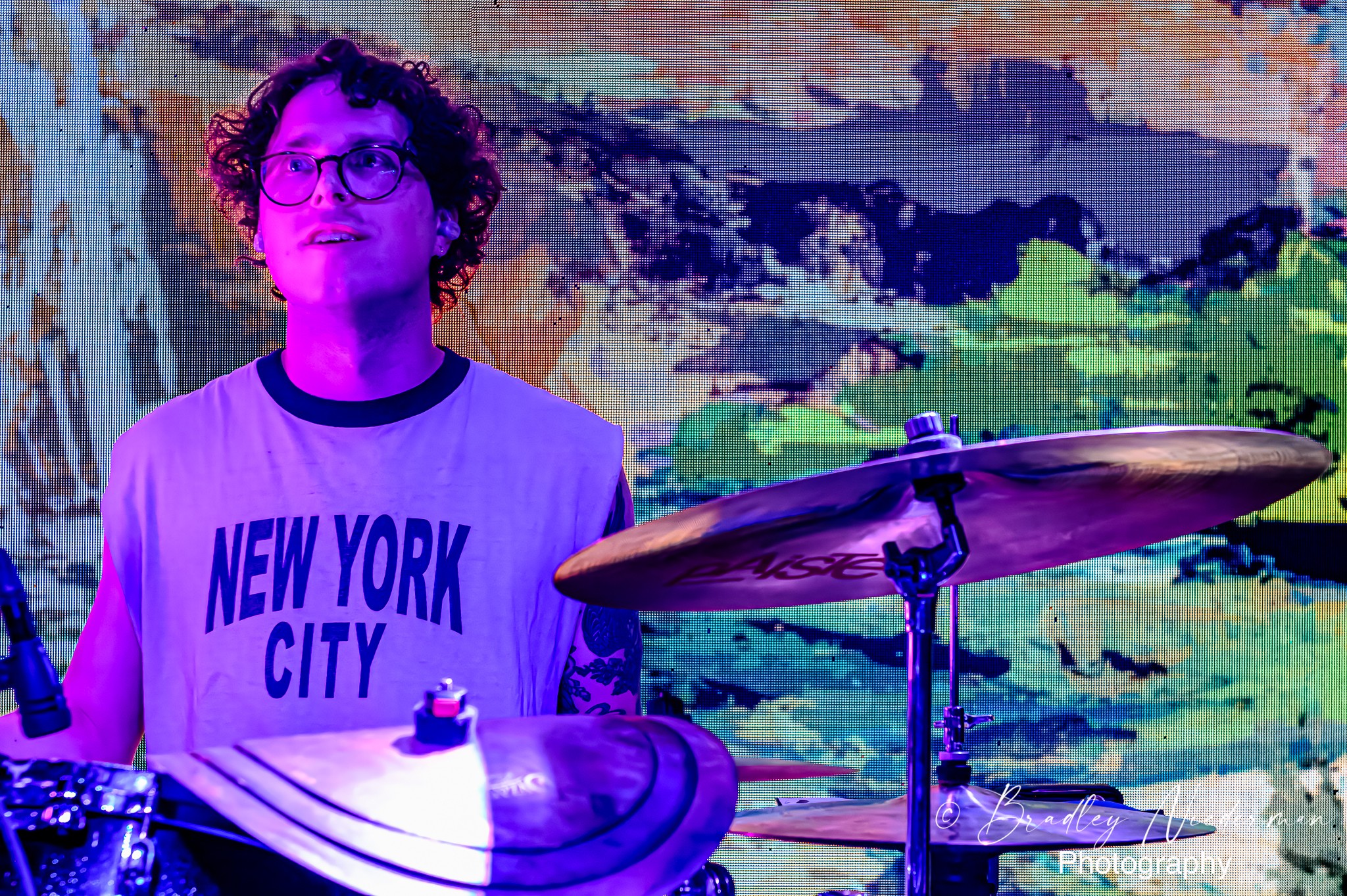Person with curly hair and glasses wearing a New York City shirt, standing near a drum set with a colorful background.