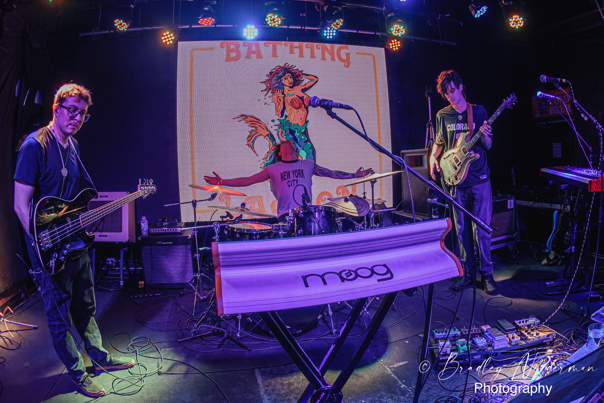 Band performs on stage with a keyboard, guitar, and drums, with a screen displaying 'BATTING' behind them.