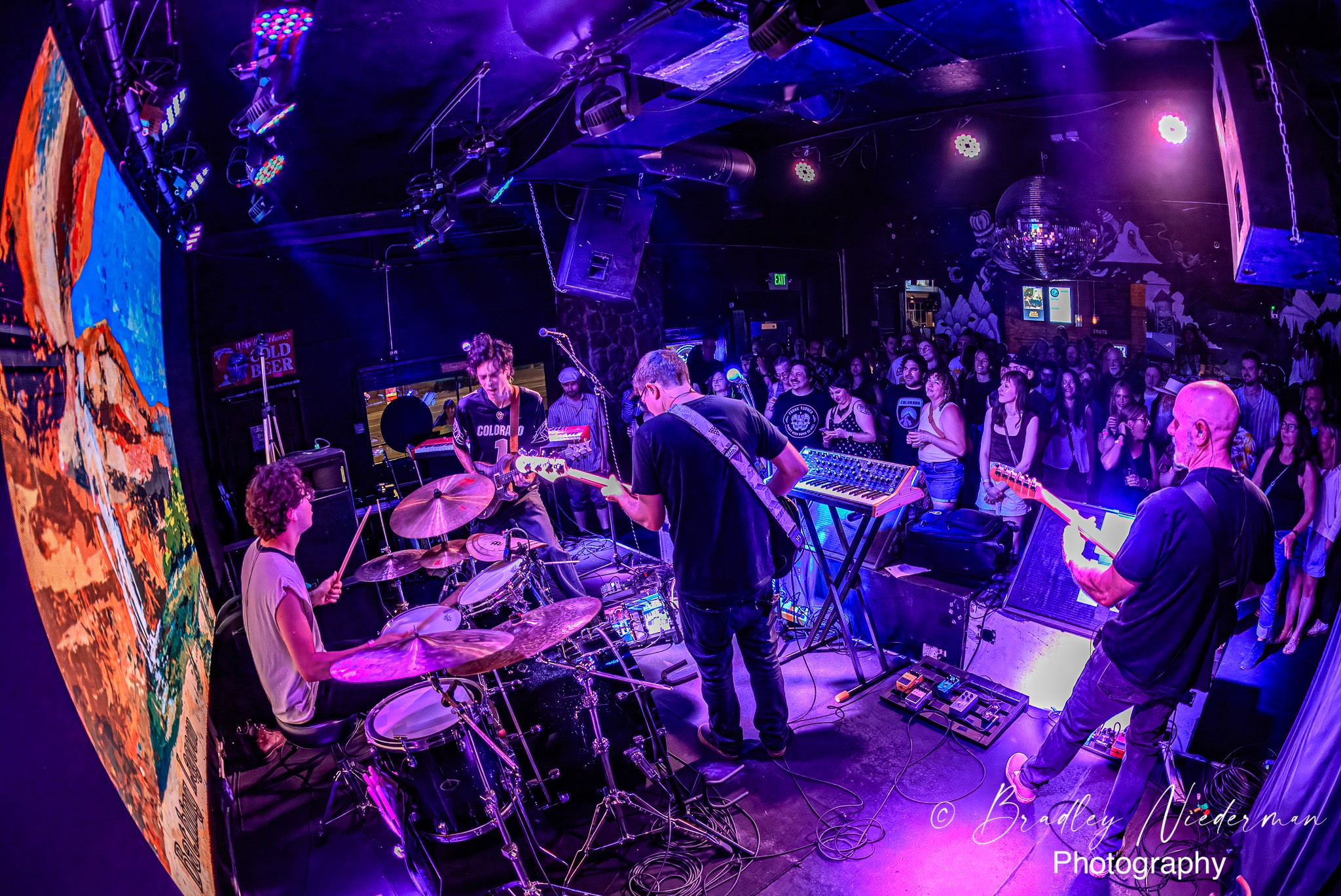 Band performs on stage with audience in a dark, colorful venue, purple and blue lighting, musical instruments visible.