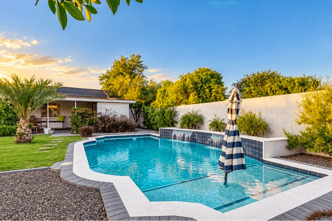 Swimming pool with waterfall feature, surrounded by greenery, a house, and a blue sky, with an umbrella nearby.
